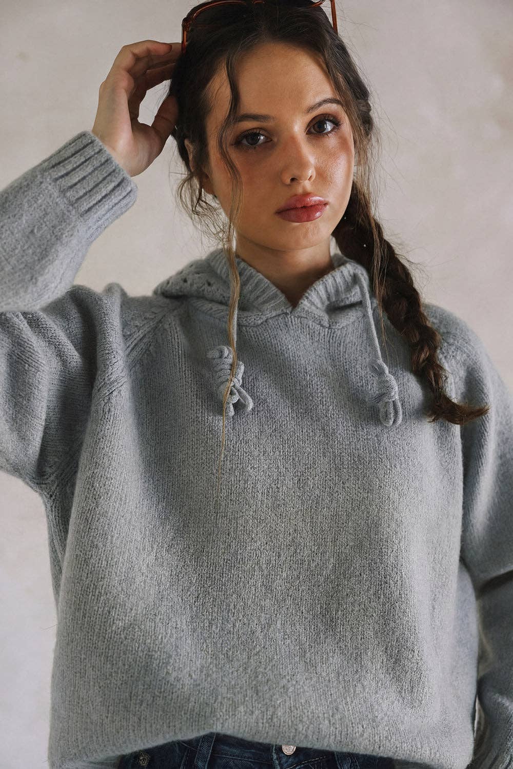 A woman wearing a light gray fuzzy knit hoodie sweater, with her hair in a side braid and sunglasses on her head, posing against a neutral light gray wall.