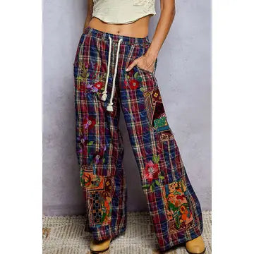 A person wearing loose, wide-leg plaid pants with colorful floral patchwork details and a white drawstring, shown from the waist down against a plain light gray wall.