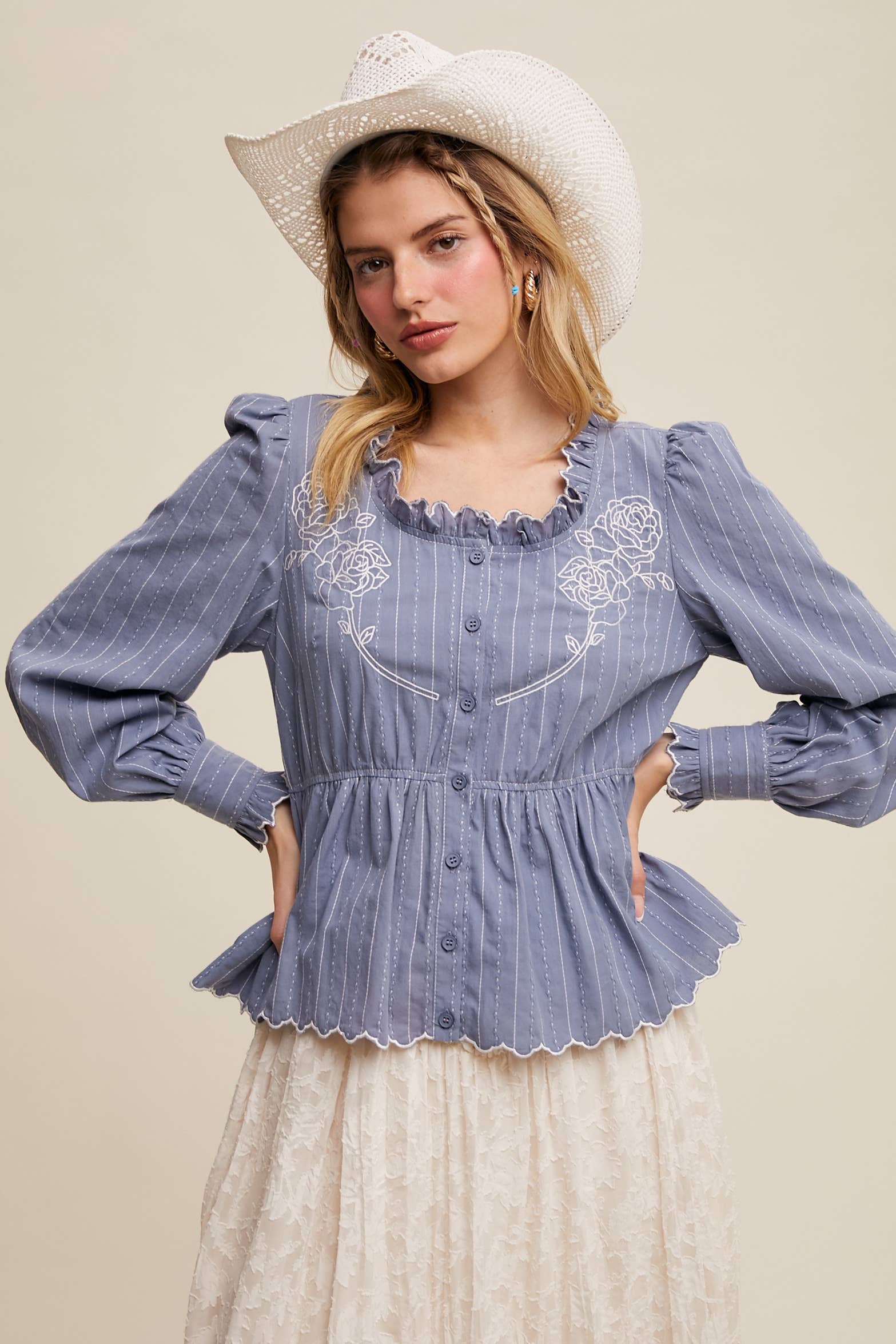 Dusty blue pinstripe blouse with ruffled neckline, white rose embroidery, puff long sleeves, button front, and scalloped peplum hem—styled with a white lace skirt and cowboy hat.