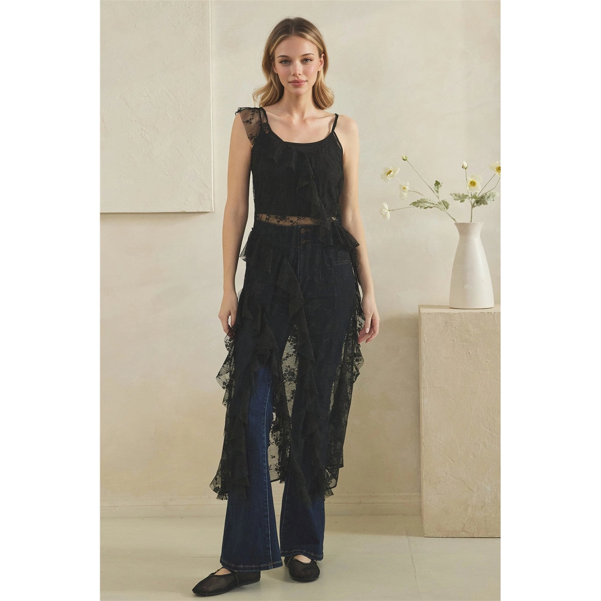 A woman models a black sheer lace duster ruffled asymmetrical hem, worn over blue jeans. She is standing in a minimalist room with a textured beige wall and a white vase on a pedestal.