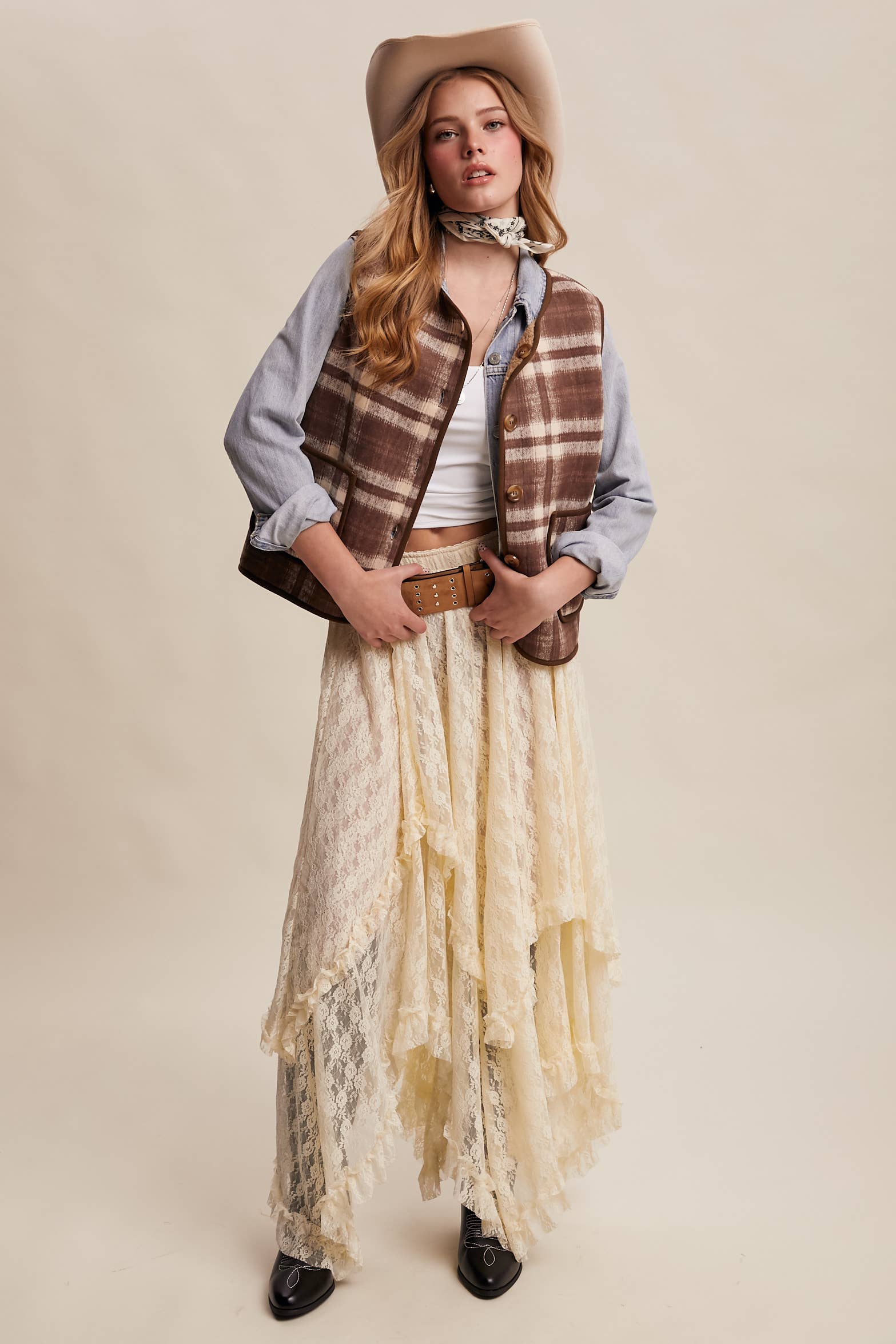 Woman wearing a plaid vest, cowboy hat, and long lace skirt on a beige background