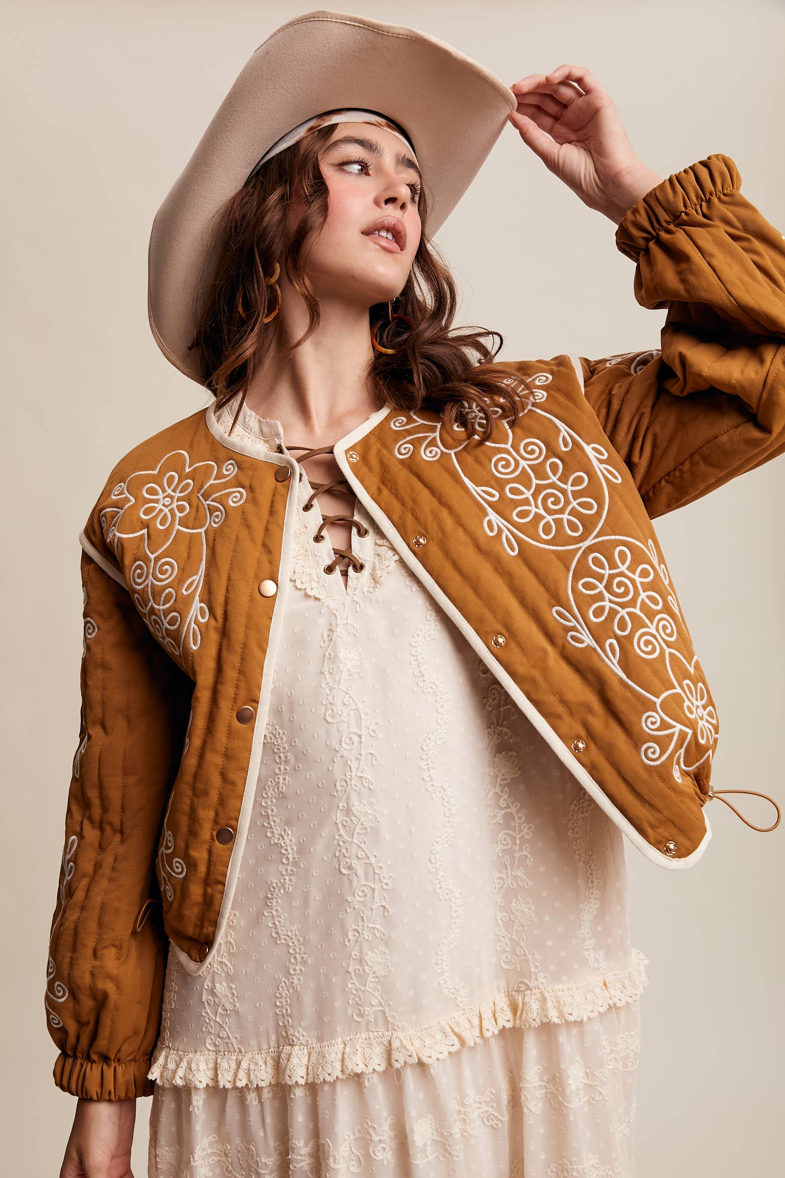 Boho caramel-brown jacket with cream floral swirl embroidery, ivory piping, and snap buttons, styled over a lace tie-front top with a scarf and felt hat outdoors.
