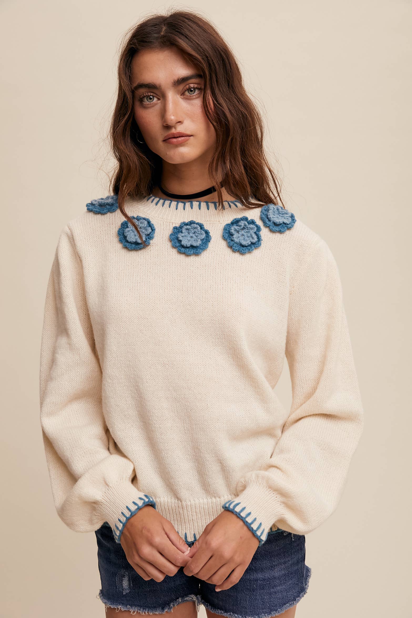 Cream sweater with blue crochet flower appliqués at neckline, blue whipstitch trim at cuffs and hem, long sleeves; styled with frayed denim shorts.
