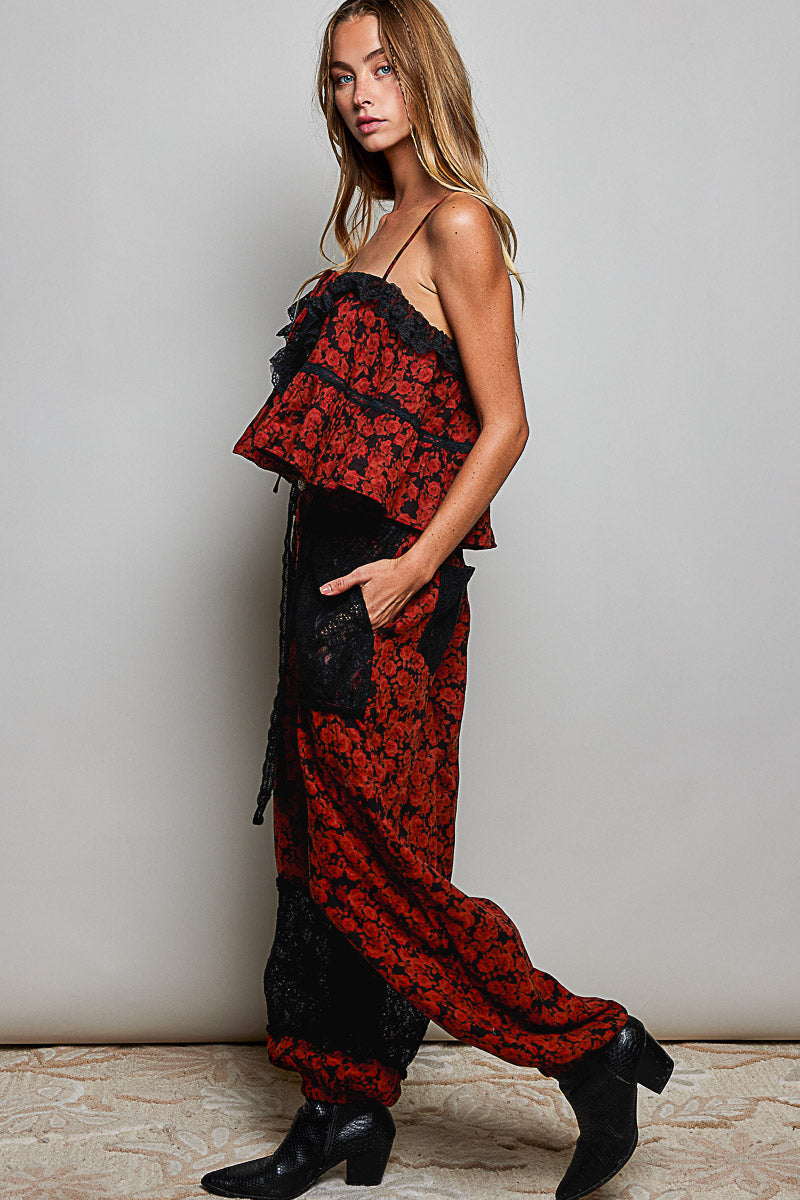 A woman modeling a red and black floral lace jumpsuit with ruffled straps and black ankle boots, standing against a light gray wall.