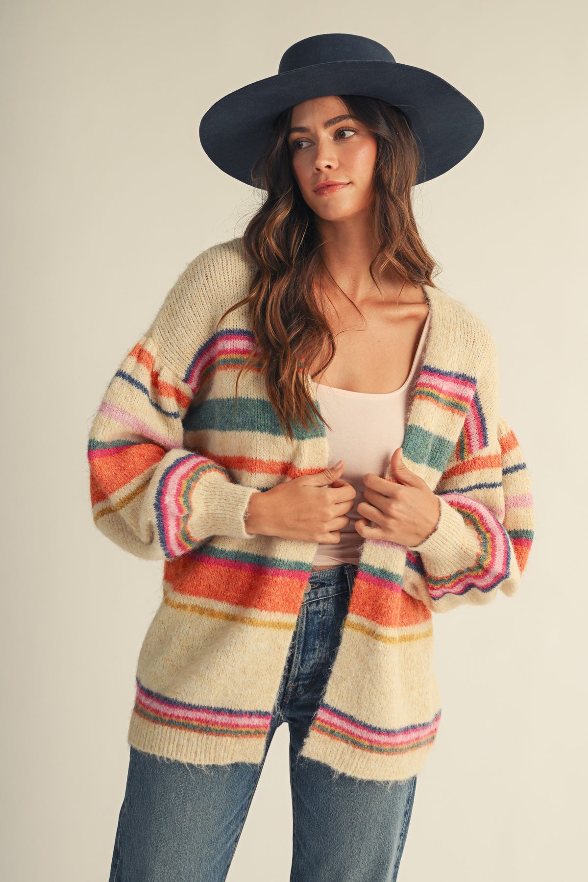 A woman wearing a cream knit cardigan with colorful horizontal stripes in pink, orange, green, and blue, paired with a black wide-brim hat, white tank top, and blue jeans, against a plain beige background.