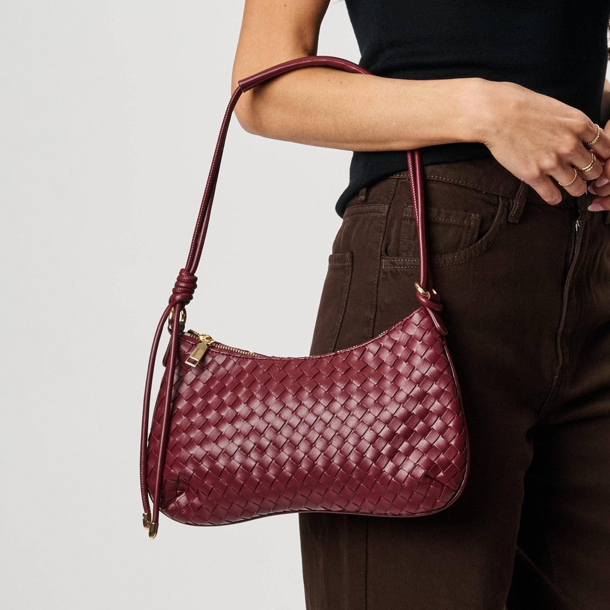 Cindy Woven Crescent Shoulder Bag in Burgundy by Moda Luxe — close-up model carry view showing woven vegan leather crescent silhouette, knotted strap detail, and gold-tone zip pull, styled with black top and denim jeans