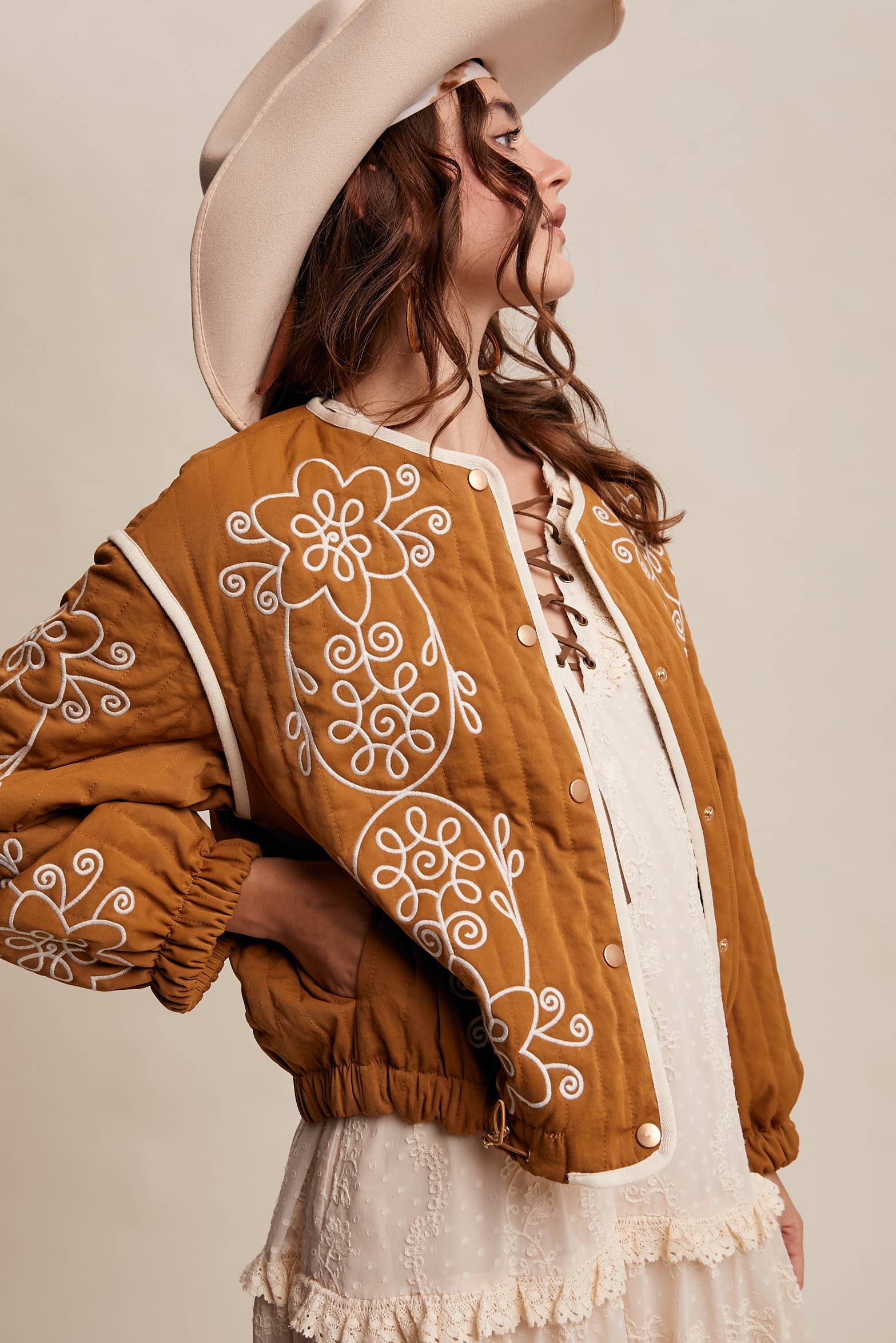 Boho caramel-brown jacket with cream floral swirl embroidery, ivory piping, and snap buttons, styled over a lace tie-front top with a scarf and felt hat outdoors.