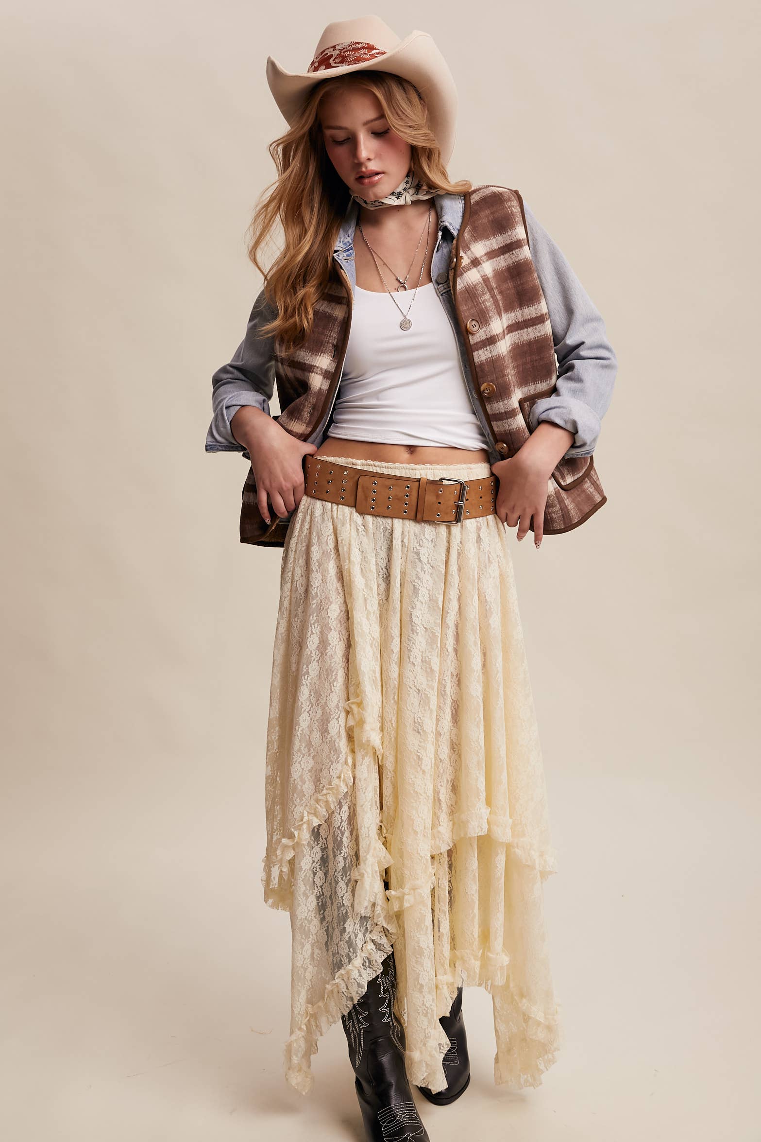 Woman wearing a cowboy hat, plaid vest, lace skirt, and boots on a beige background