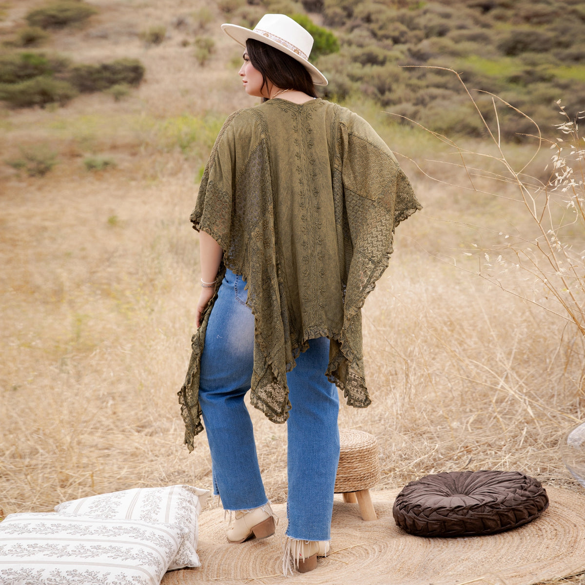 A woman stands outdoors in a dry grassy field wearing a loose olive green lace-trim poncho top, blue jeans, and a white hat, with woven floor cushions nearby.