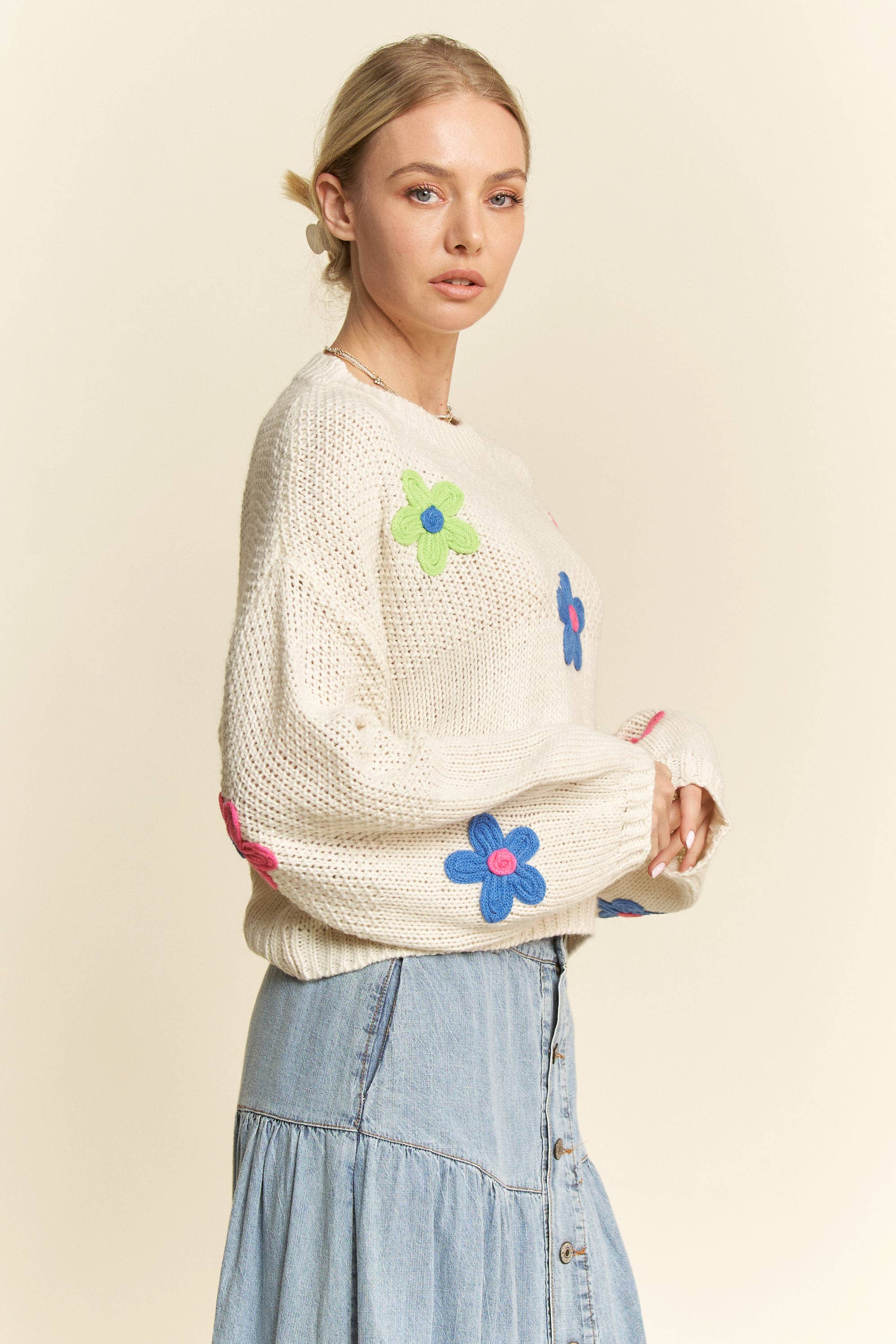 Side view- Cream knit sweater with colorful 3D floral appliqués in pink, blue, and green, styled with a button-front light denim skirt.