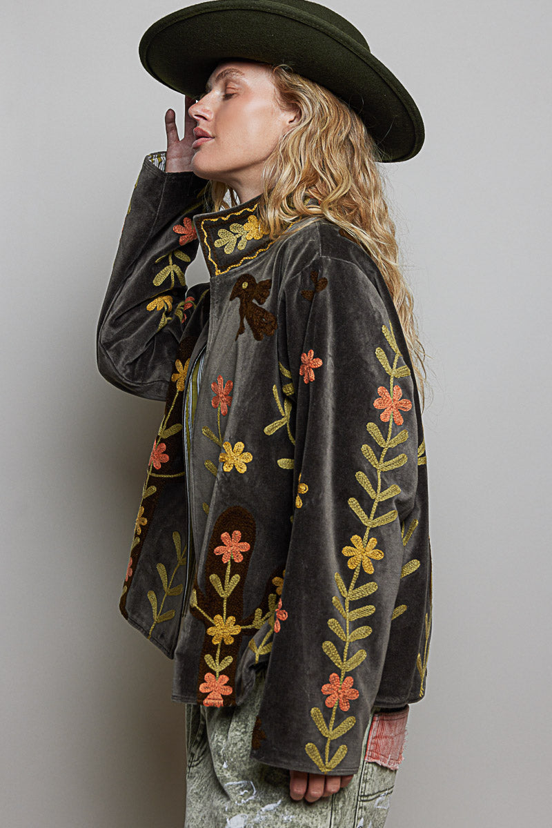 A woman wearing a dark brown velvet jacket with colorful floral embroidery, a wide-brimmed dark green hat, and light green patterned pants, posed against a neutral gray background.