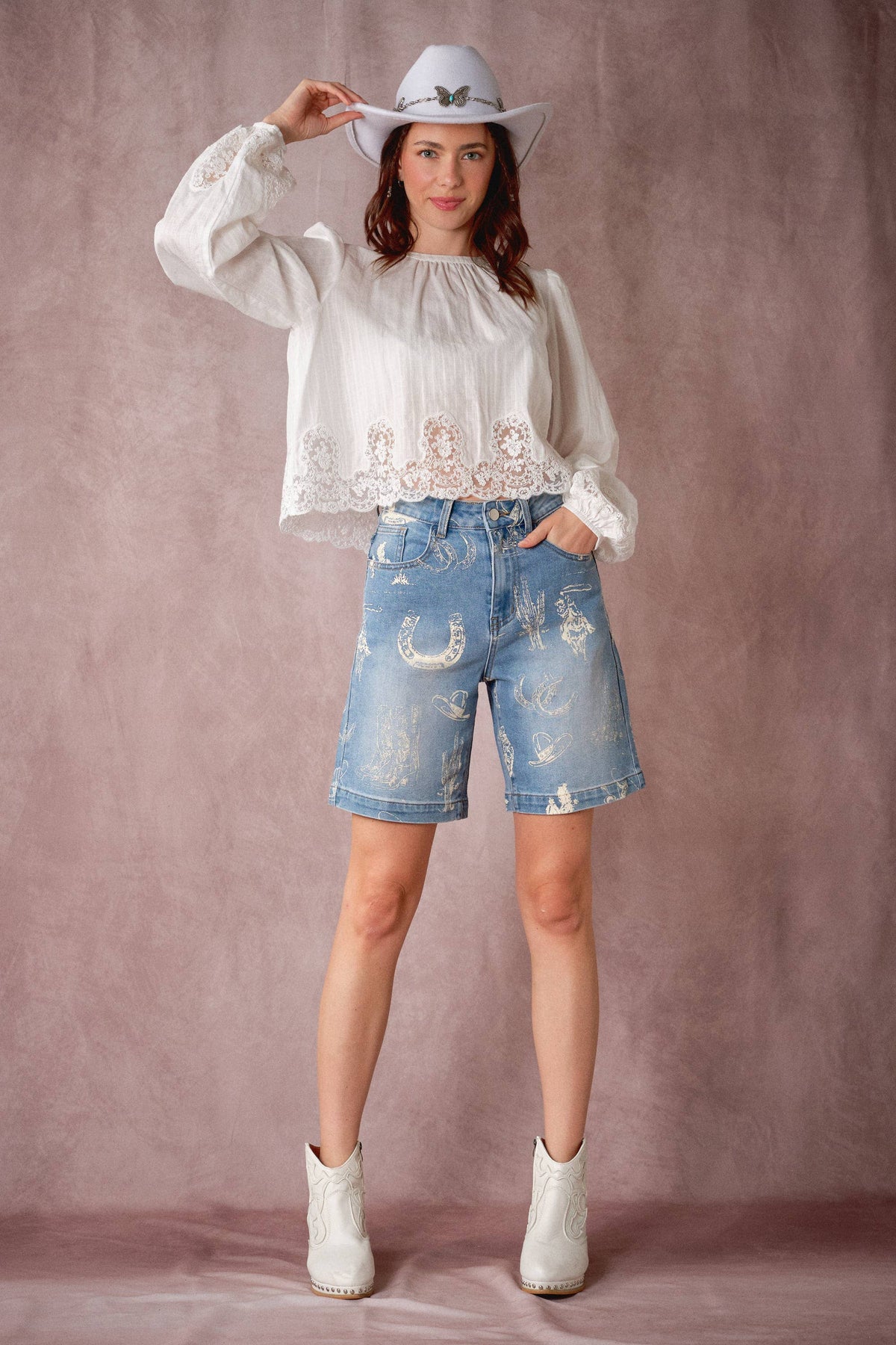 A woman modeling a western-style outfit consisting of a white long-sleeve blouse with lace trim, light-wash distressed denim shorts, a white cowboy hat, and white ankle boots, posed against a dusty mauve studio backdrop.