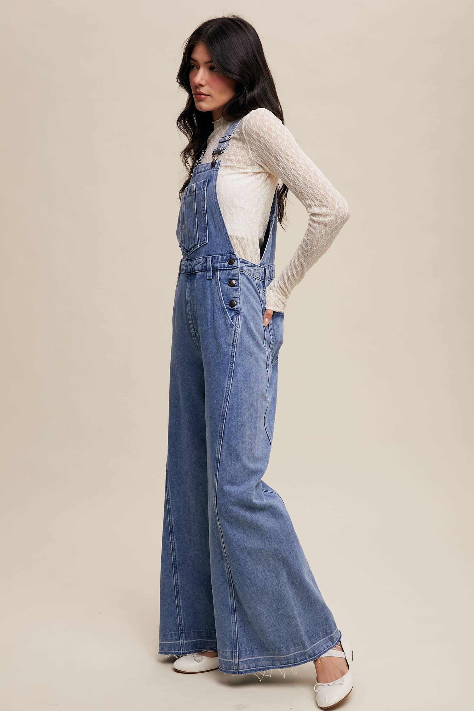 Side view- Light-wash wide-leg denim overalls with adjustable straps, front bib pocket, side buttons, and slant pockets, finished with a raw hem and layered over a cream long-sleeve top.