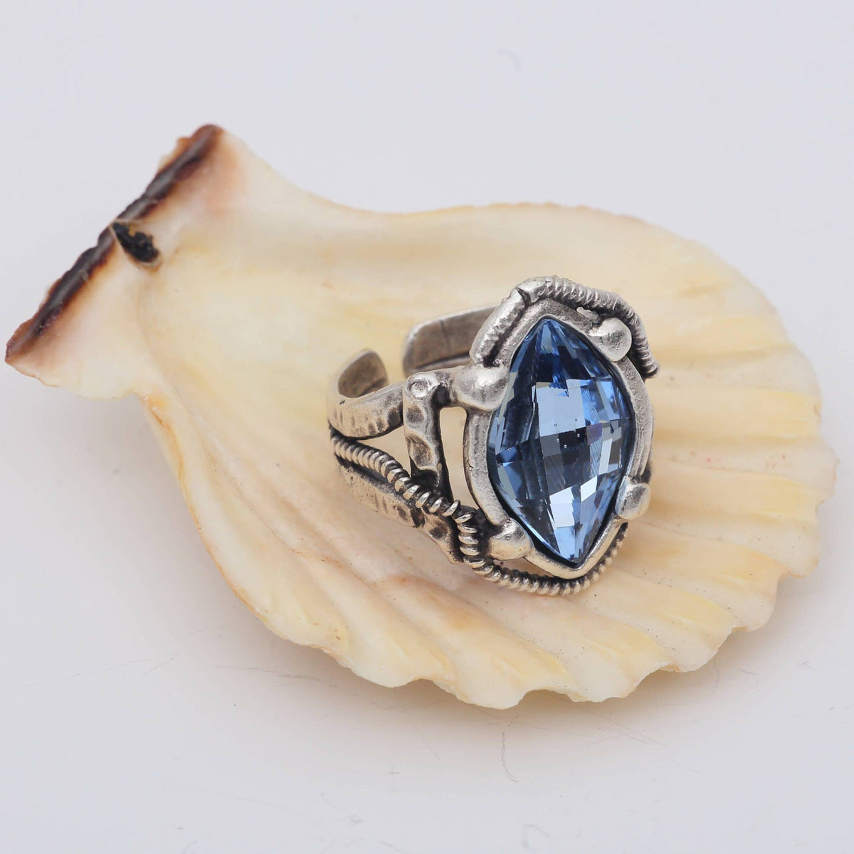 A silver-toned ring with a marquise-cut blue gemstone, displayed on a beige scallop shell against a white background.