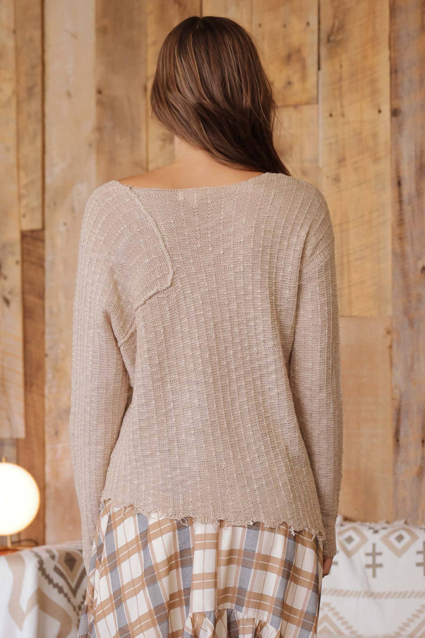 Back view of model wearing taupe distressed lightweight sweater with ribbed knit texture, raw edge hem, and relaxed fit paired with tan plaid ruffle skirt