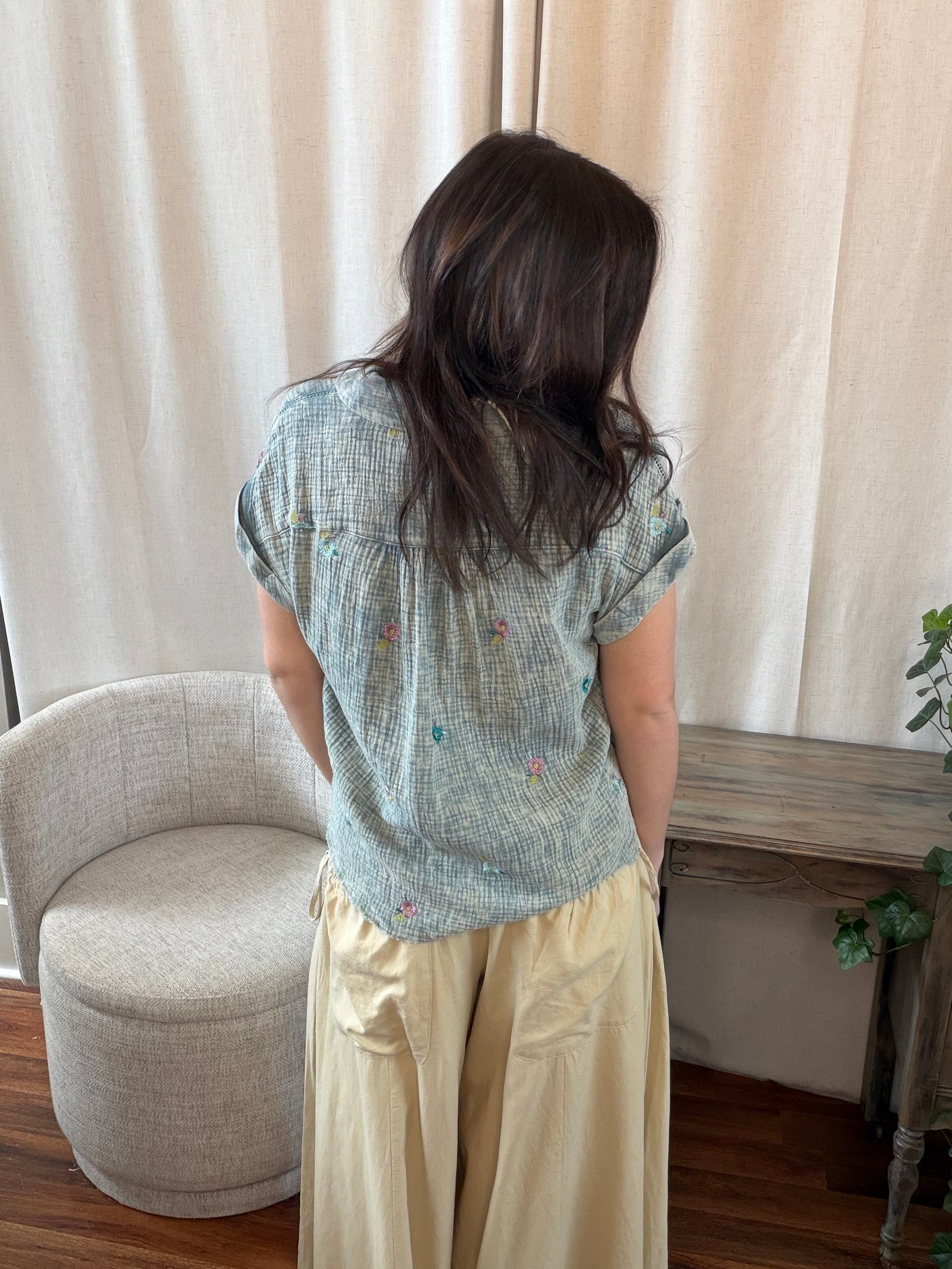 Person wearing a light blue patterned shirt and beige pants standing in a room with curtains and a chair.