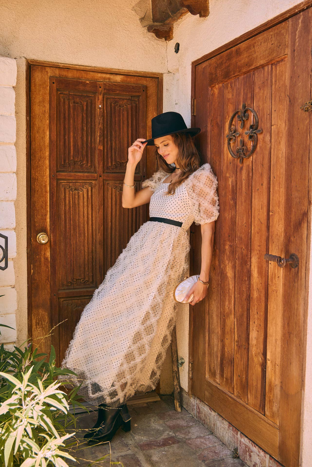 eaning pose — cream polka-dot tulle midi dress with puff sleeves and black belt, styled with black wide-brim hat and ankle boots by a wooden door.