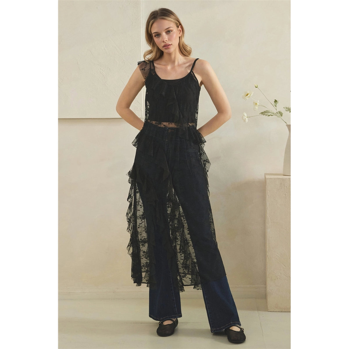 A woman modeling a black sheer lace sleeveless duser with ruffles, styled with black flats in a neutral indoor setting.