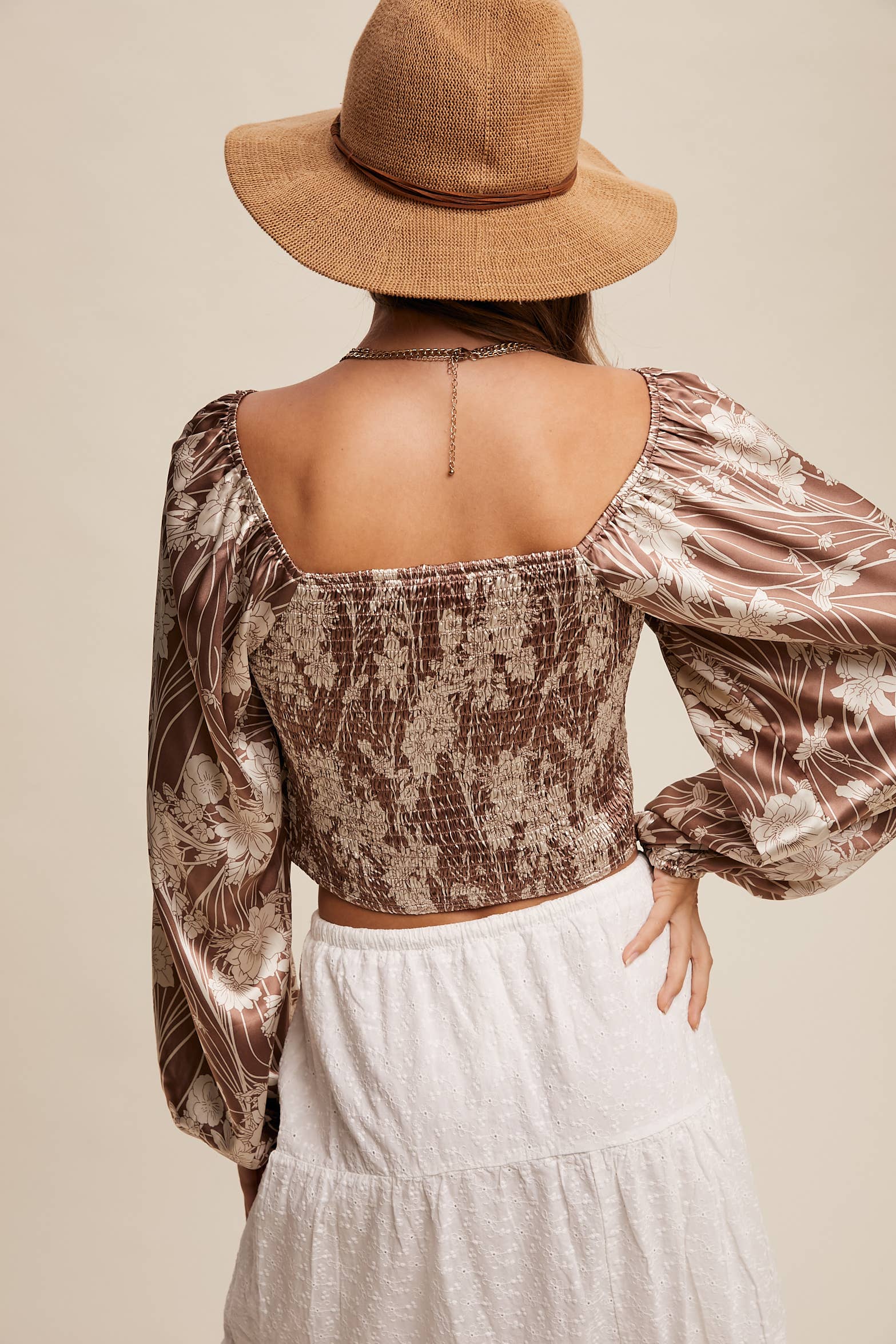 Back view- Brown and cream floral smocked blouse with square neckline, puffed long sleeves, and elastic cuffs, styled with layered necklaces and a wide-brim hat.
