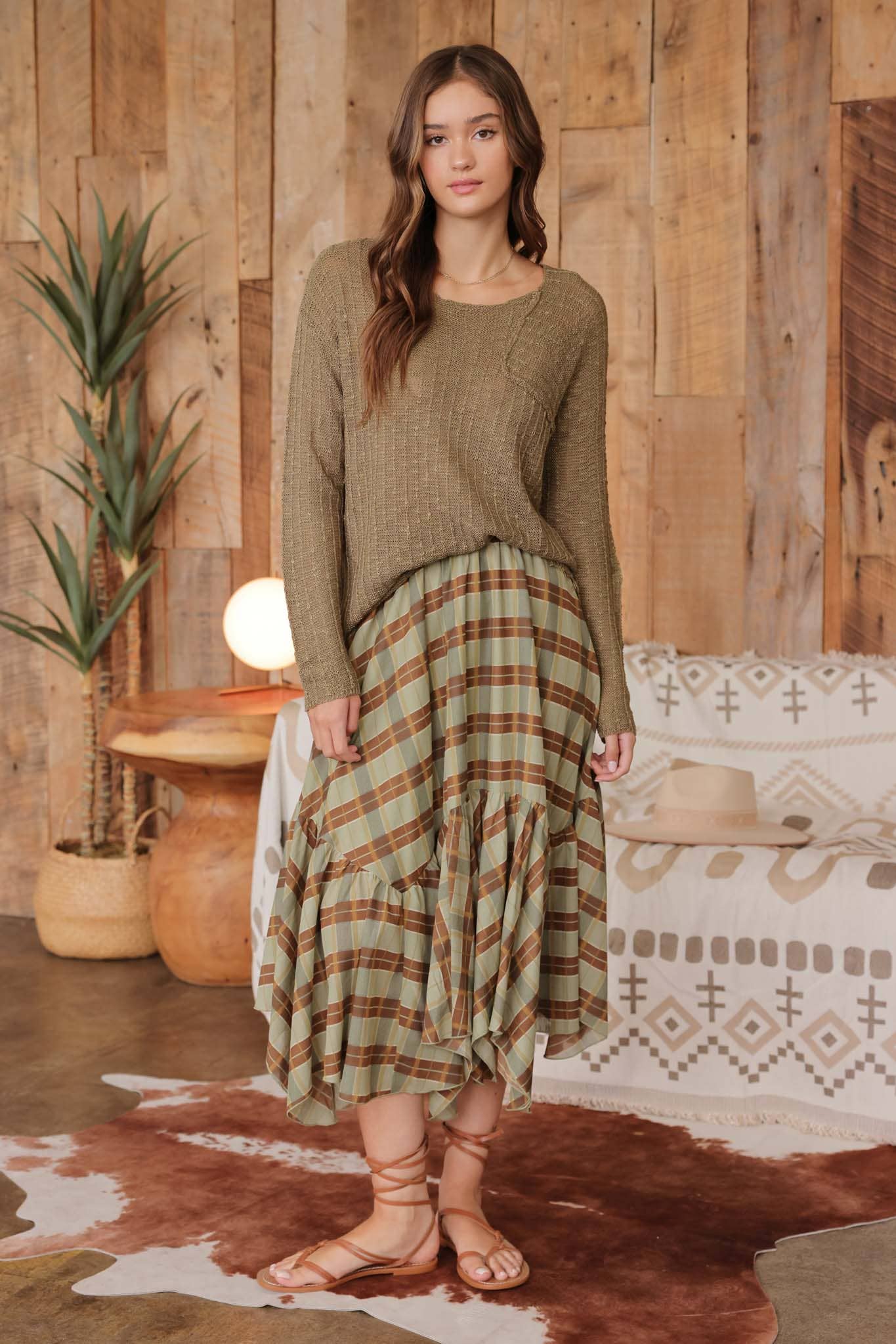 Front view  of model wearing olive distressed lightweight sweater with ribbed knit texture and raw edge hem, paired with a green and brown plaid ruffle skirt and lace-up sandals.