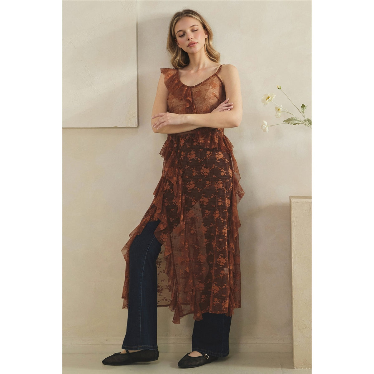 A woman models a copper sheer floral lace duster with a ruffled tiered skirt, styled over dark blue jeans and black flat shoes against a neutral beige wall.