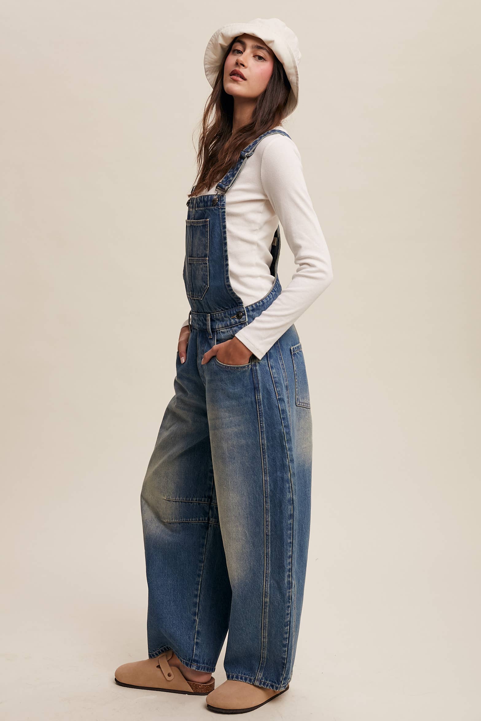 Side view- Vintage-inspired blue denim overalls with relaxed wide-leg fit, front chest pocket, and adjustable straps, styled with a white long-sleeve top and neutral clogs.