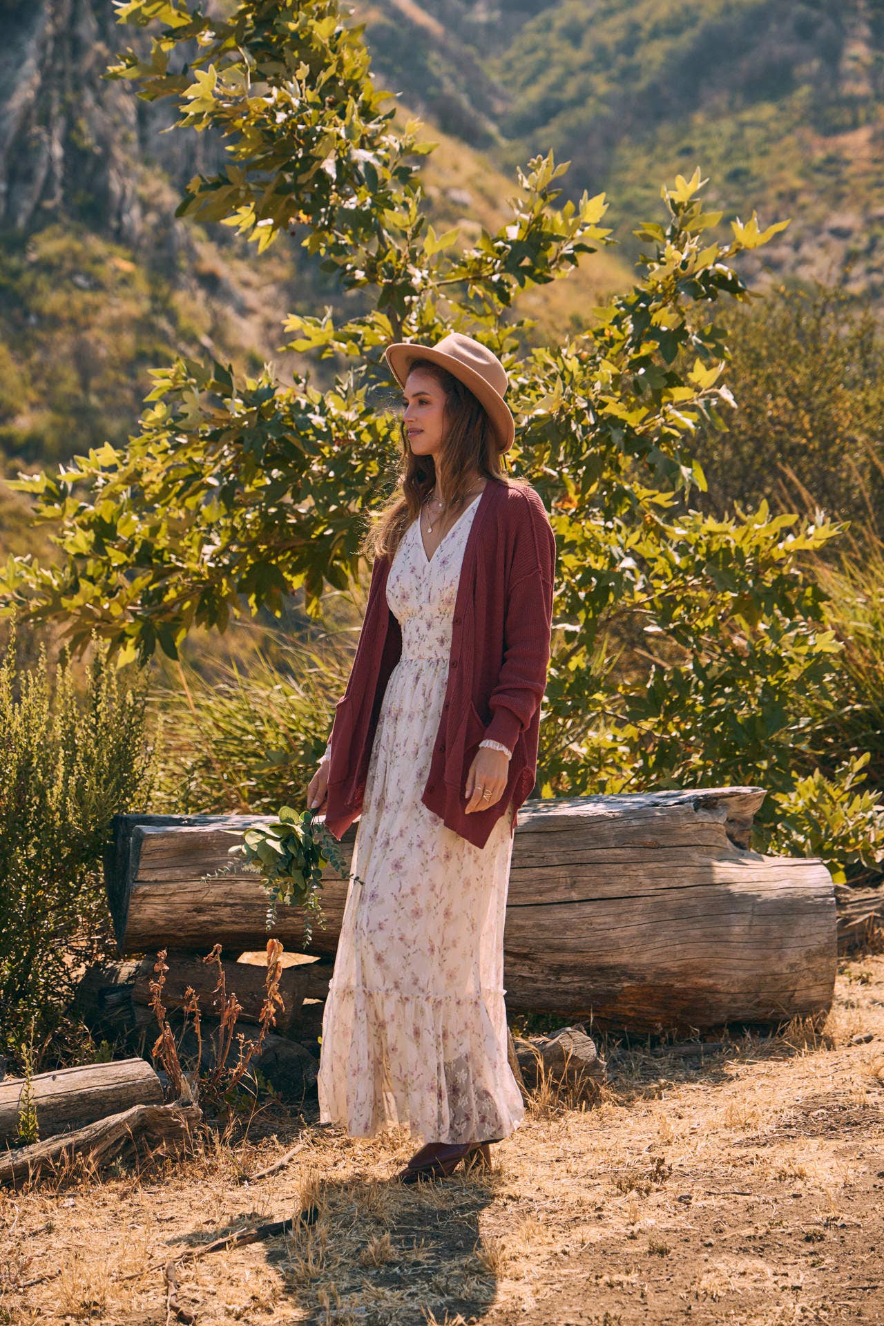 Front view — cream floral maxi dress with smocked waist and ruffle hem; mauve cardigan, tan hat, holding a greenery bouquet outdoors.