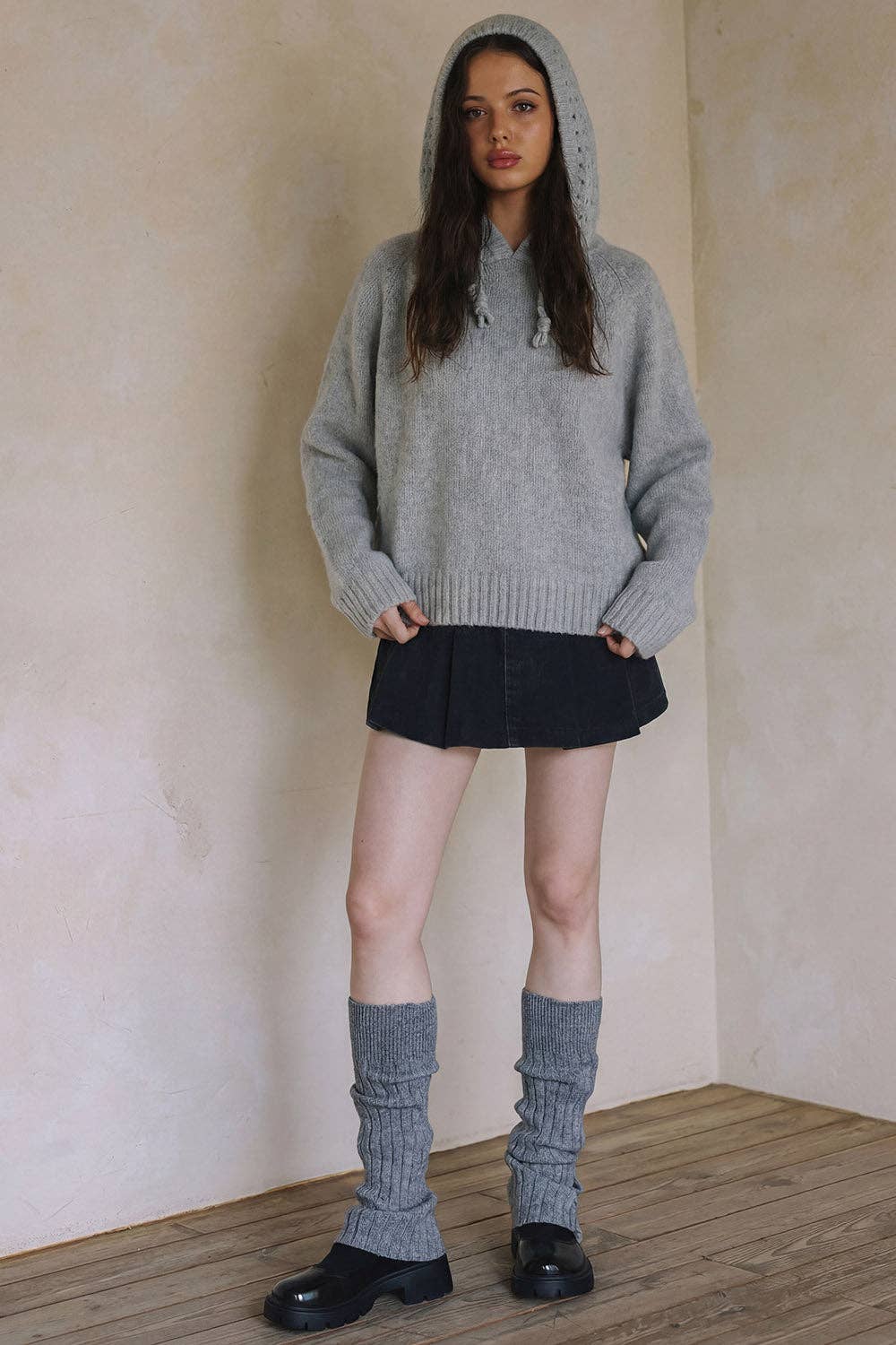 A woman stands in a textured beige room wearing a light gray oversized knit hoodie, a black mini skirt, gray leg warmers, and black shoes.
