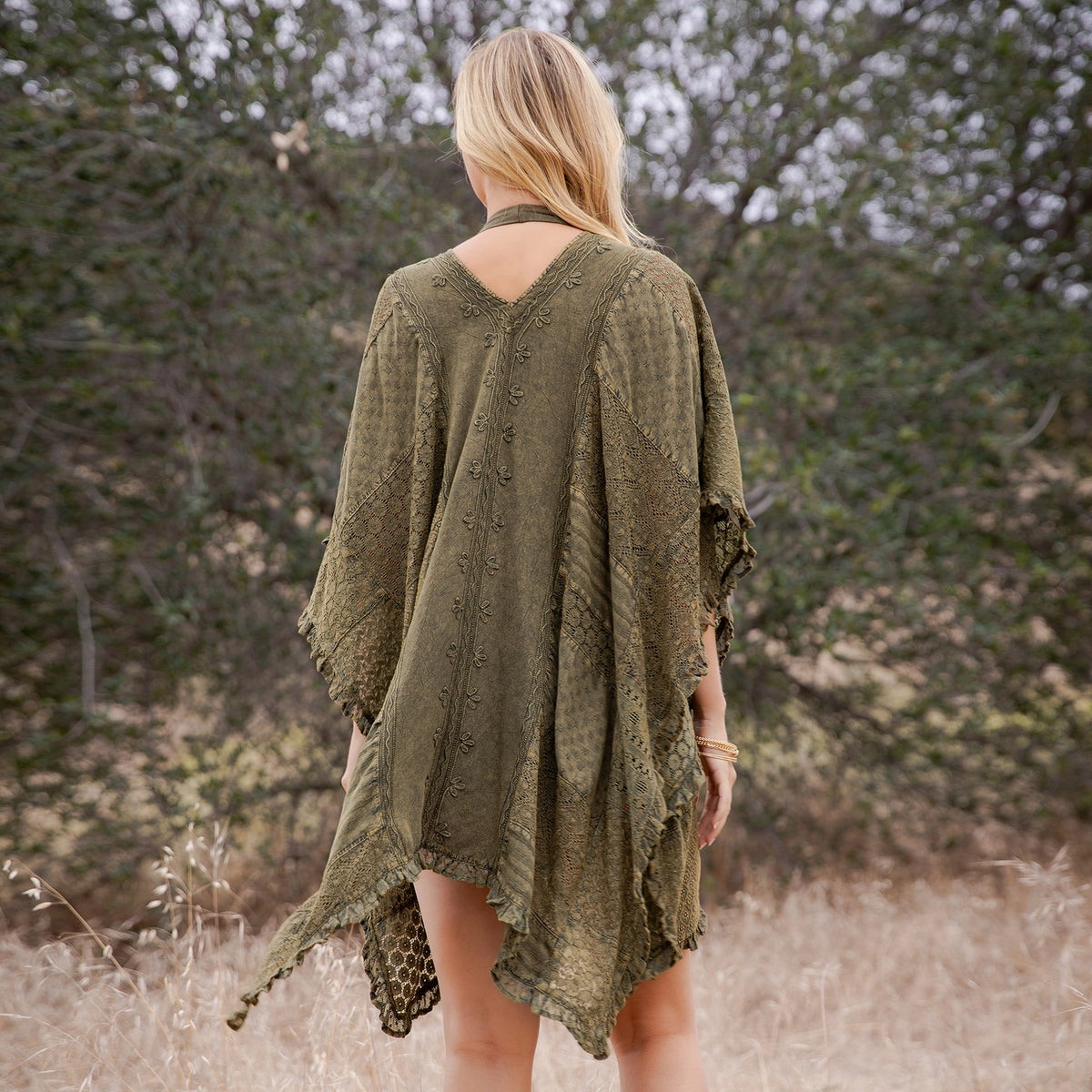 A woman viewed from behind wearing an olive green textured crochet poncho cover-up in an outdoor field with trees in the background.