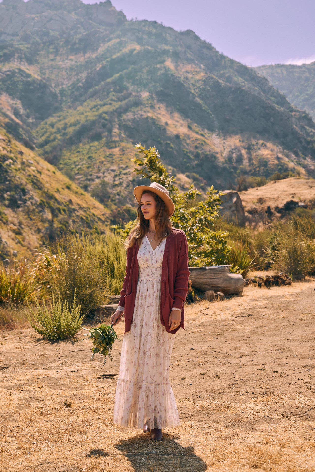 Front view — cream floral maxi dress with smocked waist and ruffle hem; mauve cardigan, tan hat, holding a greenery bouquet outdoors.