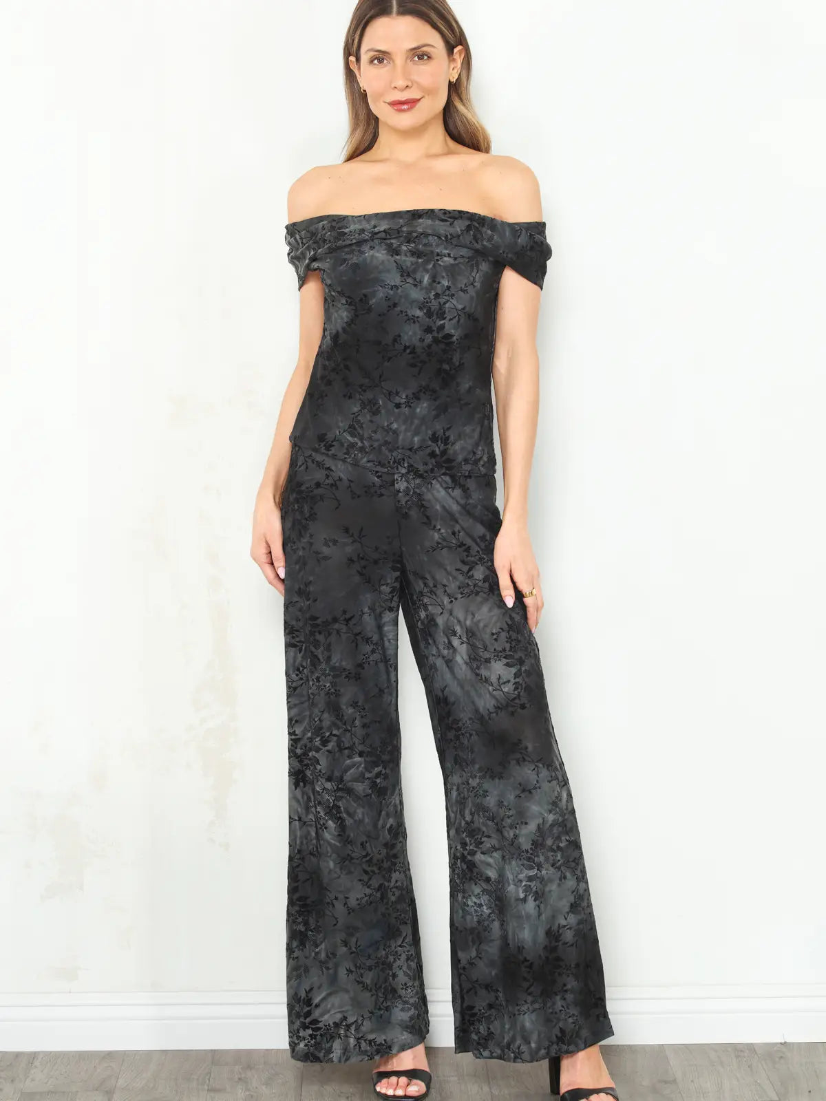 A woman modeling an off-the-shoulder black jumpsuit with a subtle tonal floral jacquard pattern, set against a plain white wall.