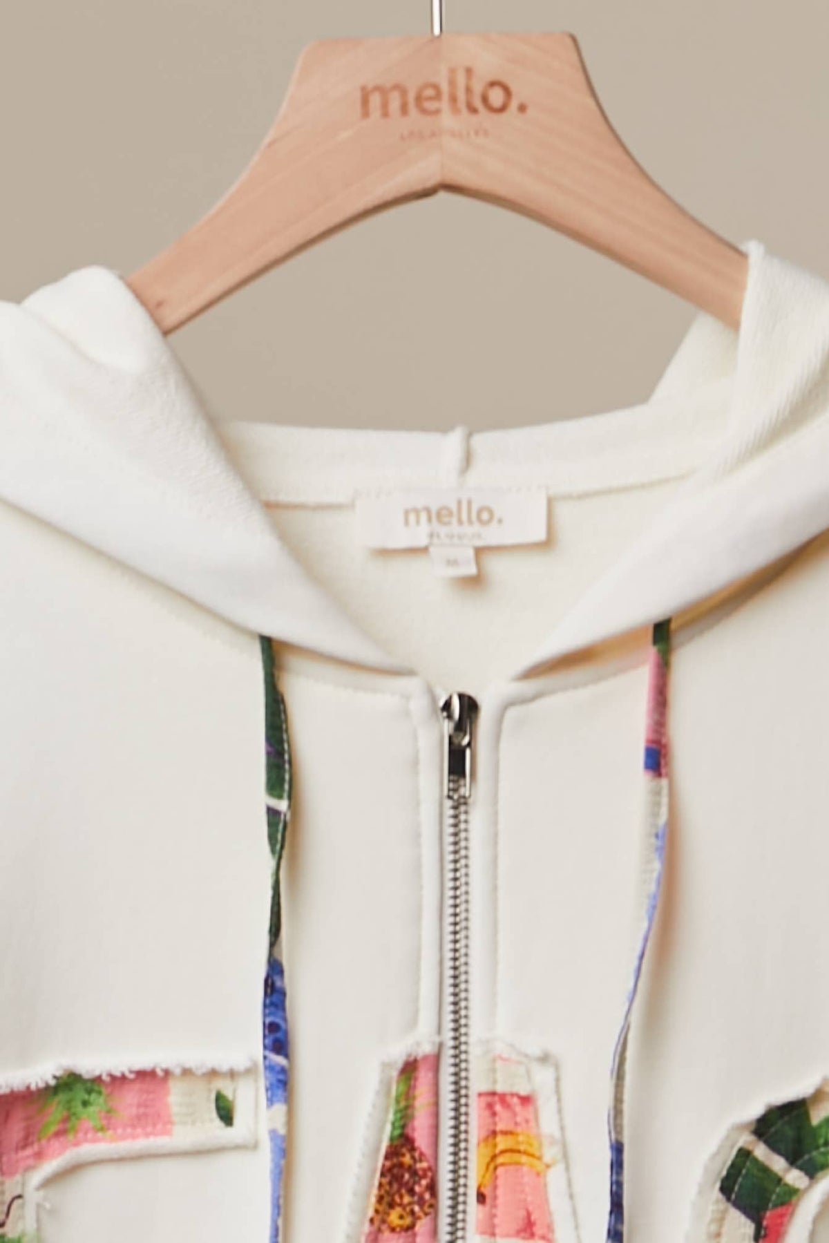 A close-up of a cream-colored zip-up hoodie with colorful patchwork details on the pockets and drawstrings, hanging on a light wood hanger branded with 'mello.'.