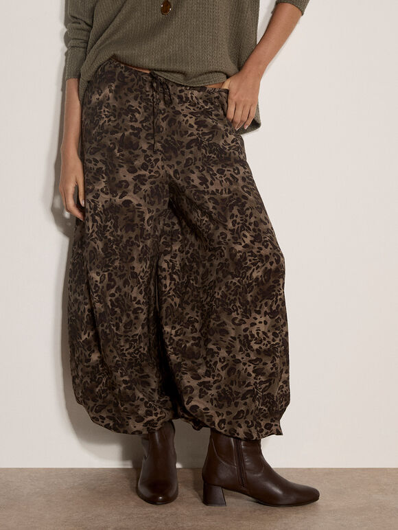 A person wearing loose, balloon-style brown pants with an allover leopard print, paired with a muted green top and dark brown ankle boots, standing against a plain off-white wall.