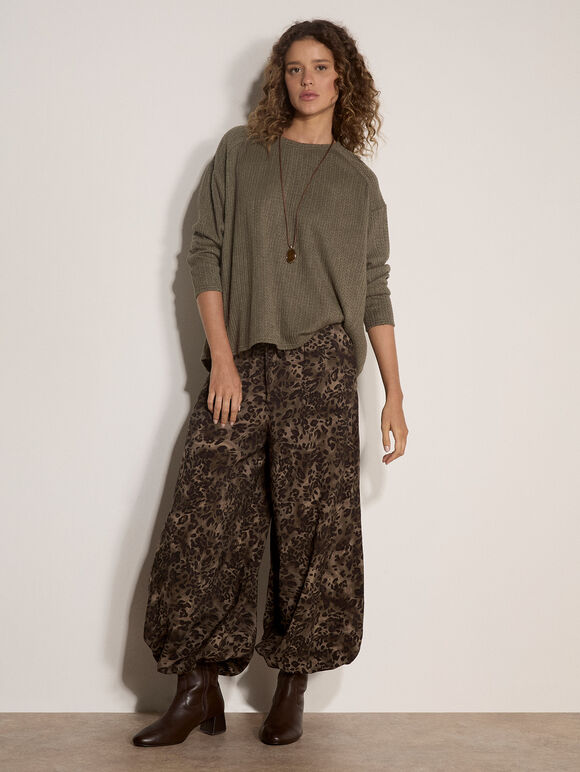A woman stands against a plain off-white wall wearing a loose olive green long-sleeve top, brown patterned wide-leg pants, and dark brown ankle boots.