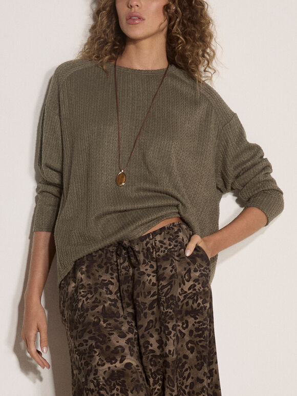 A woman wearing an olive green ribbed knit long-sleeve sweater and brown leopard-print pants, accessorized with a long pendant necklace, against a plain off-white background.