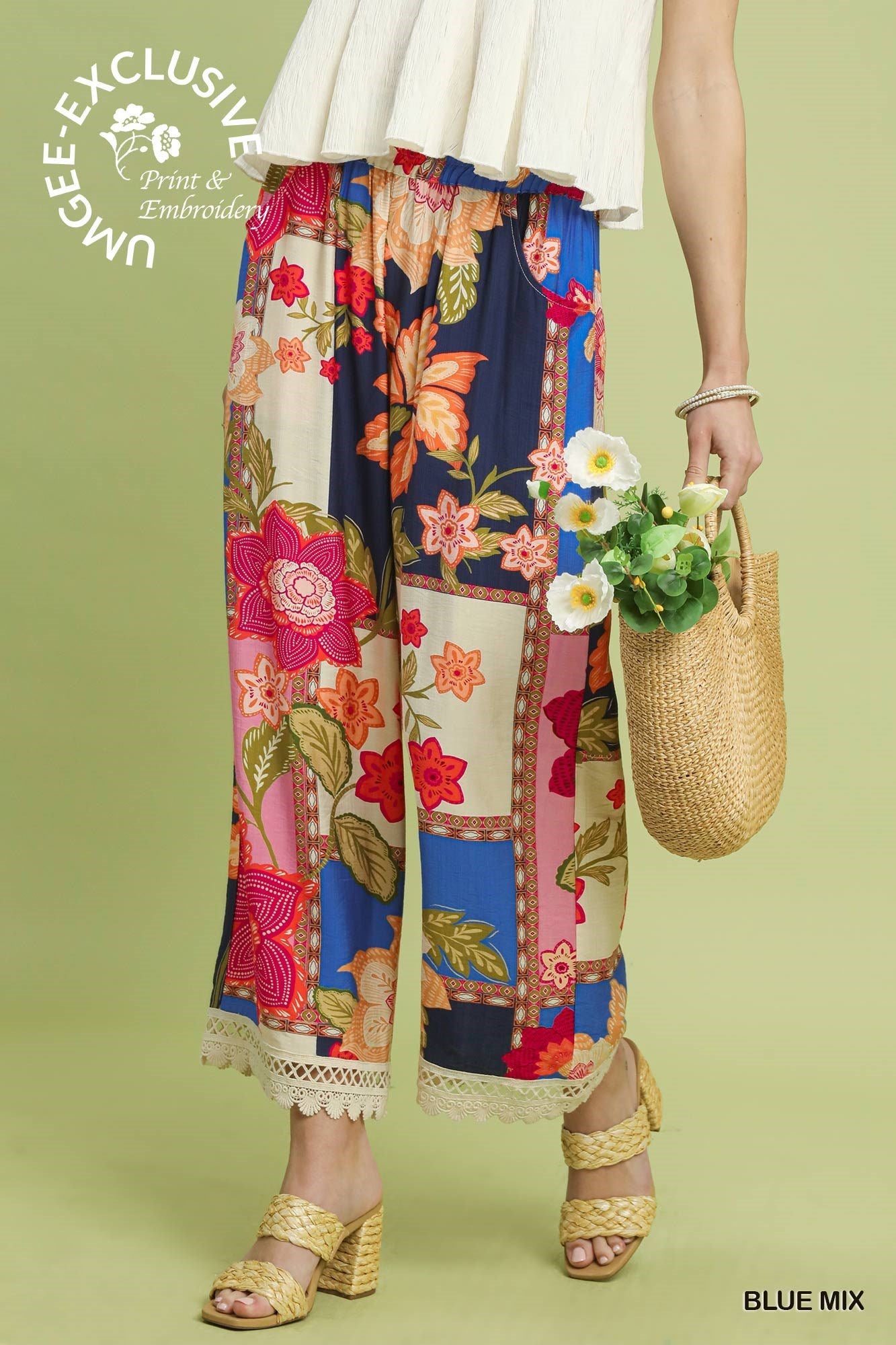 Woman wearing colorful floral pants with a white top on a green background, holding a woven bag.