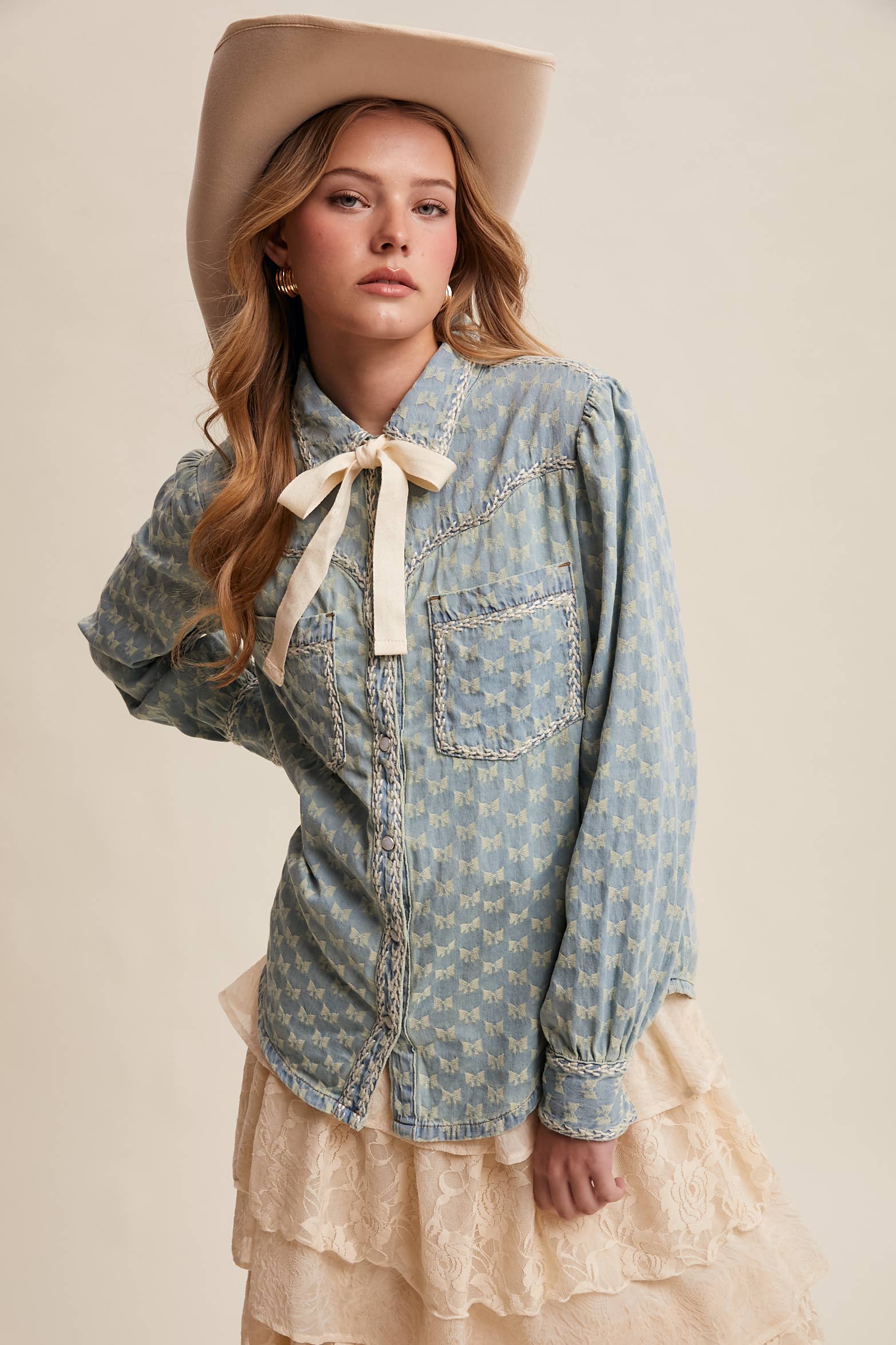 Light-wash denim blouse with cream bow-tie collar, tonal mini-bow pattern, braided contrast stitching, snap front, and two chest pockets.
