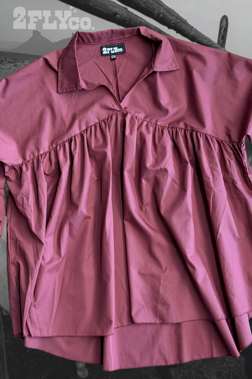 A dusty rose pink short-sleeve collared shirt by 2FLYCo, featuring a gathered waist seam and a button-down front, laid flat on a dark wooden surface.