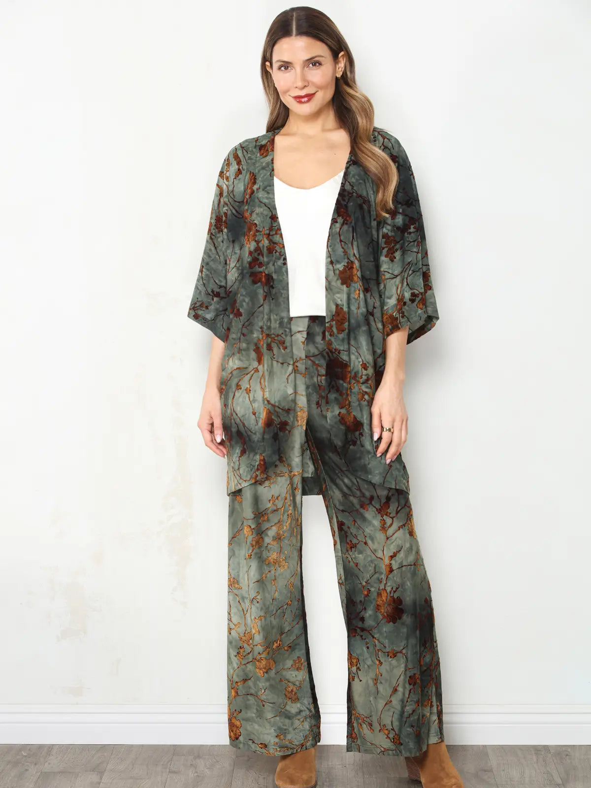 A woman modeling a matching two-piece lounge set featuring a kimono-style cardigan and wide-leg pants in a muted green and brown abstract floral print, worn over a white top.