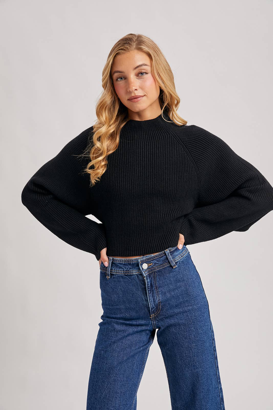 Black ribbed mock-neck sweater with long wide sleeves and a cropped fit, styled with high-rise blue jeans.