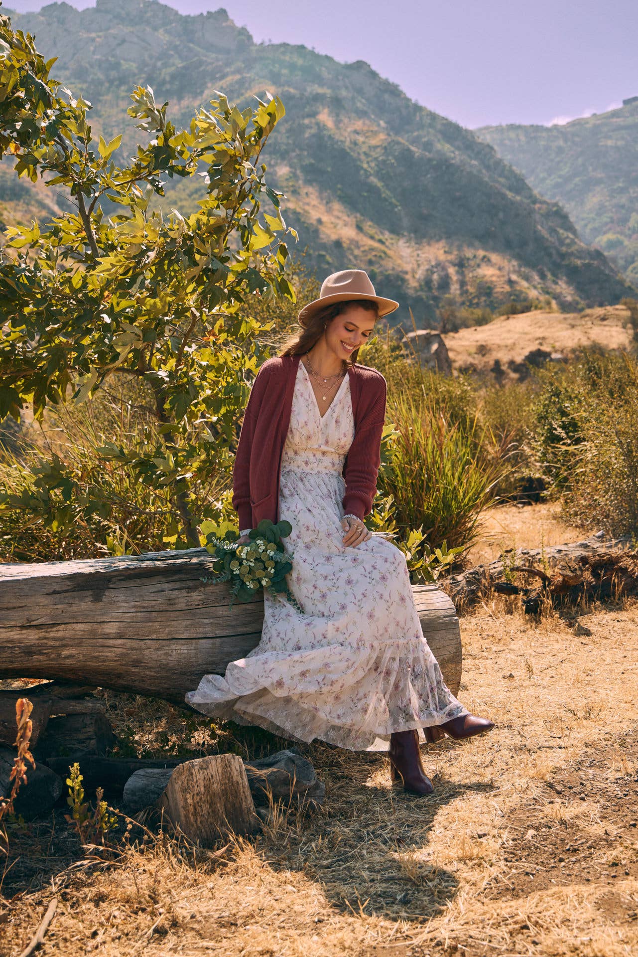 Seated view — cream floral maxi dress with smocked waist and ruffle hem; mauve cardigan, tan hat, ankle boots, holding bouquet.