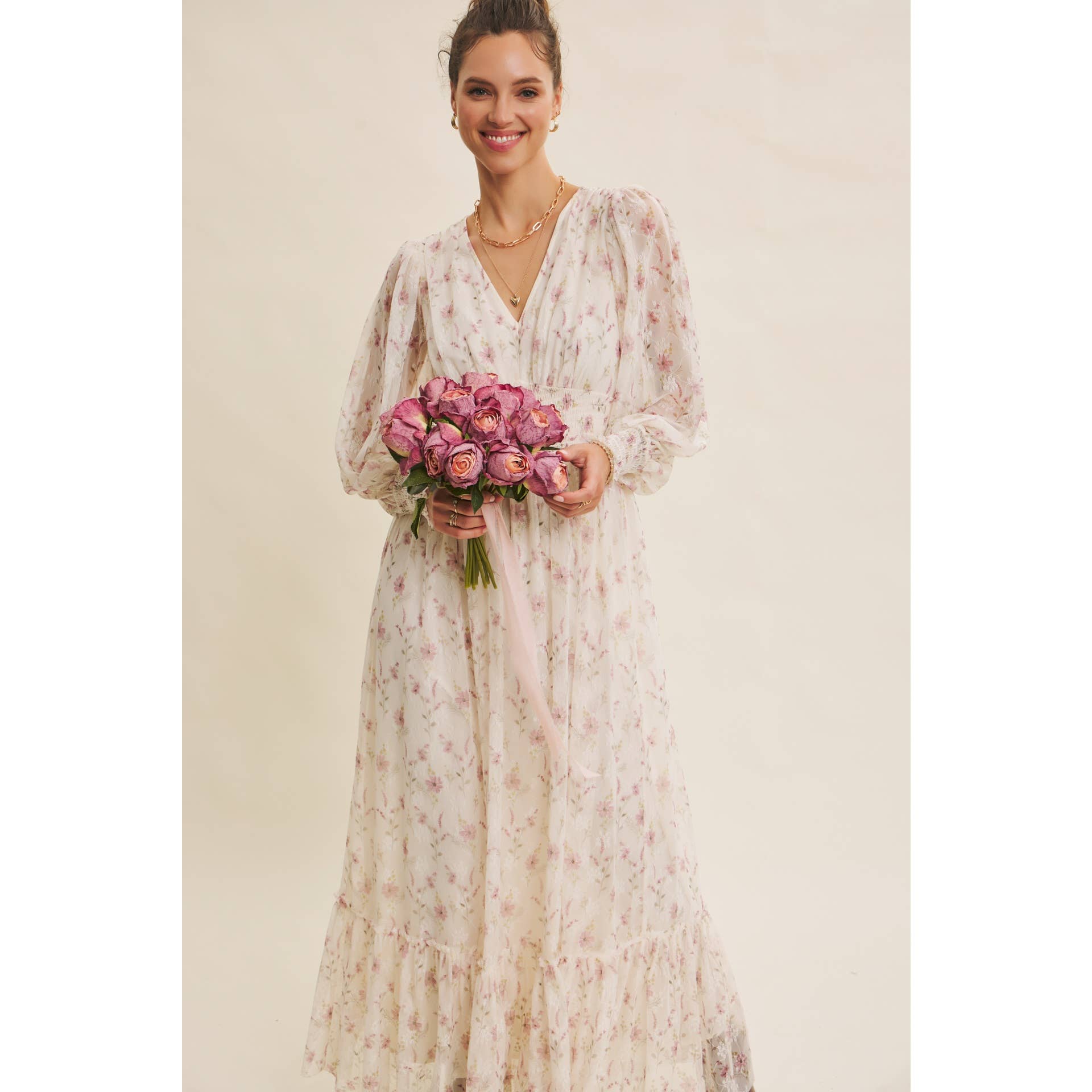 Front view — cream floral chiffon maxi dress with V-neck, sheer long sleeves, smocked waist, ruffle hem; model holding pink bouquet.