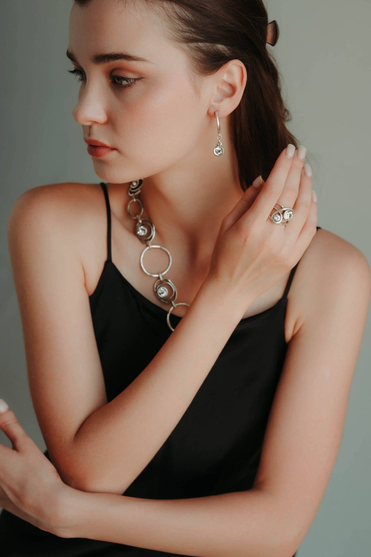 A woman wearing a silver-toned multi-ring necklace, matching drop earrings, and a ring with clear stones, styled with a black camisole against a soft gray background.