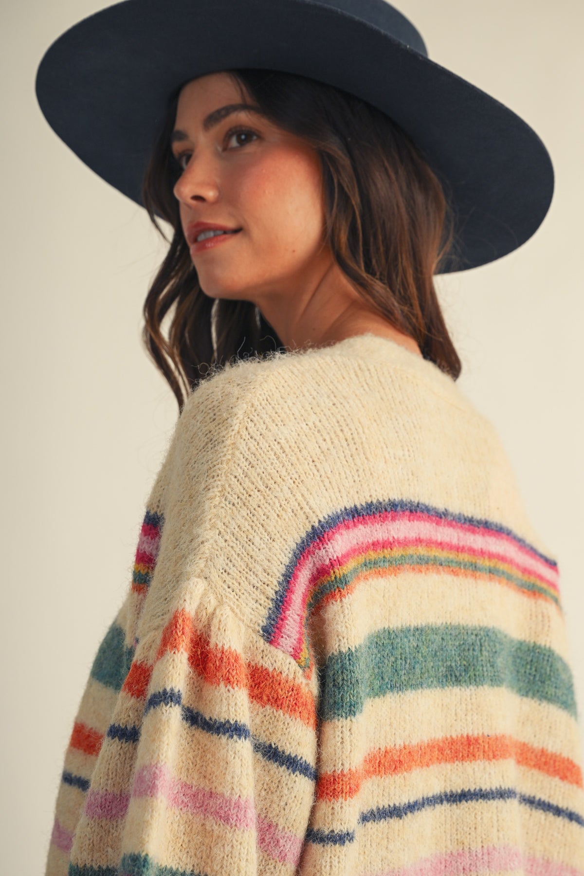 A woman wearing a cream knit sweater with colorful horizontal stripes in pink, orange, green, and blue, paired with a dark navy wide-brim hat, shown from a side-back angle against a plain light beige background.
