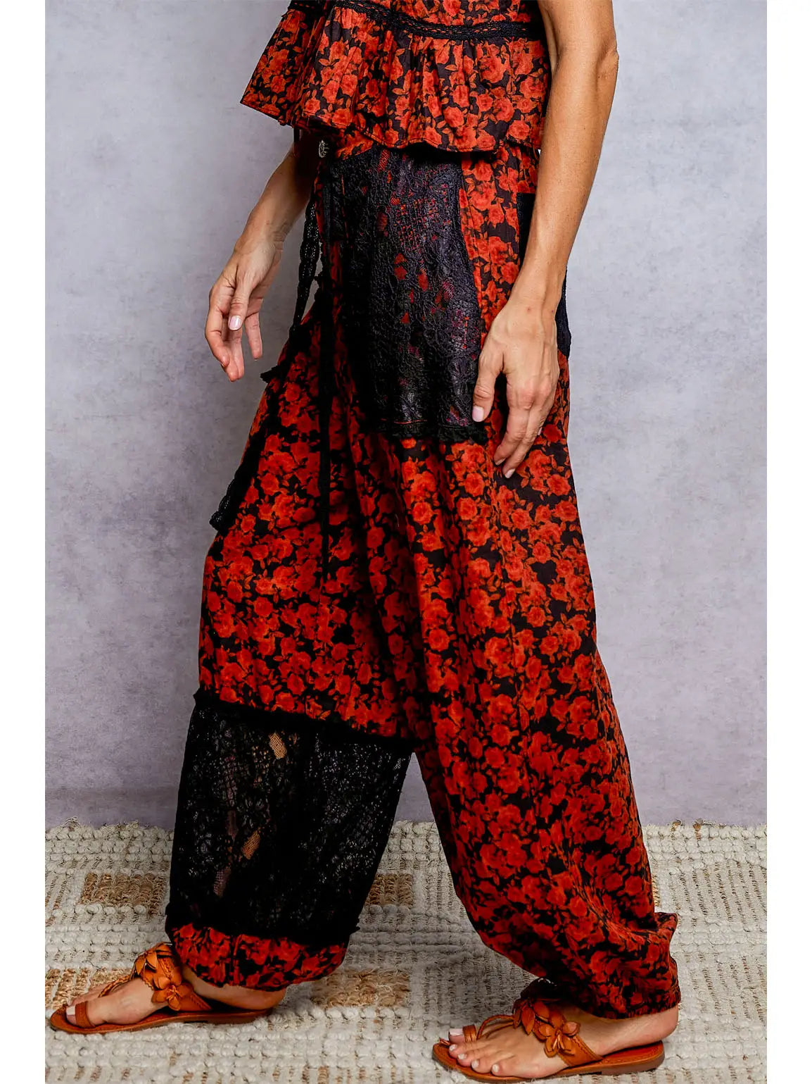 A person wearing loose harem-style pants with a red and black floral print, sheer black lace side panels, and brown strappy sandals, standing against a gray textured backdrop.
