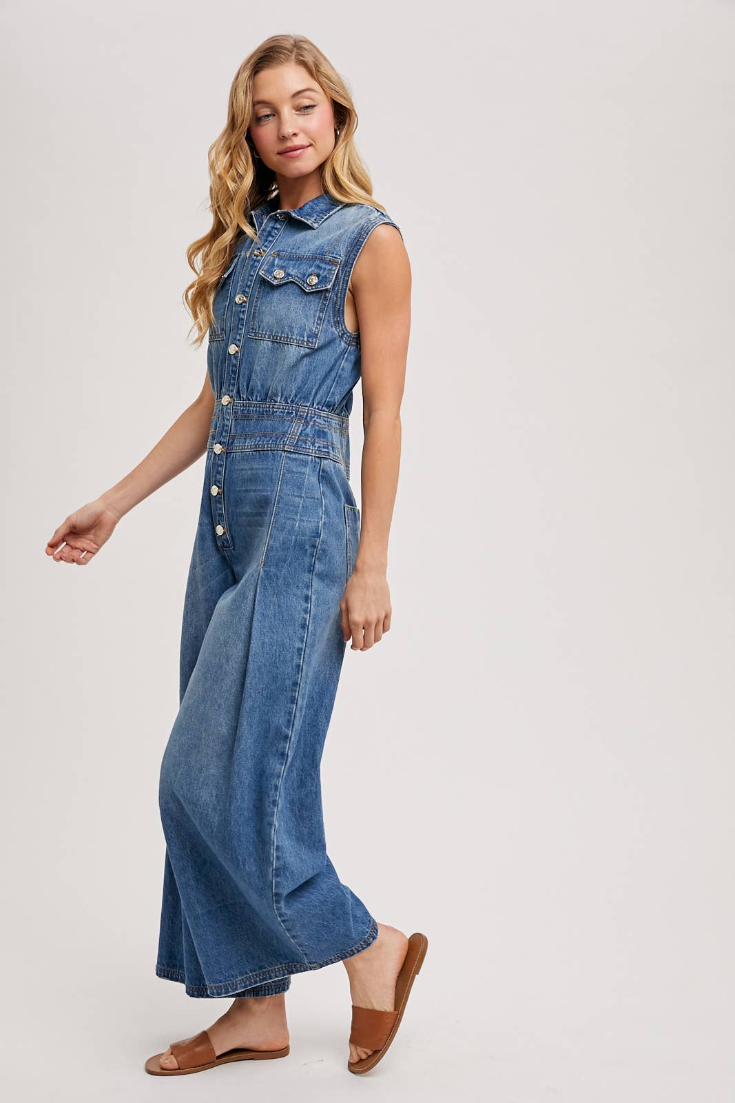 Side view of sleeveless denim jumpsuit with collared button-front and wide-leg legs, worn with brown slide sandals.