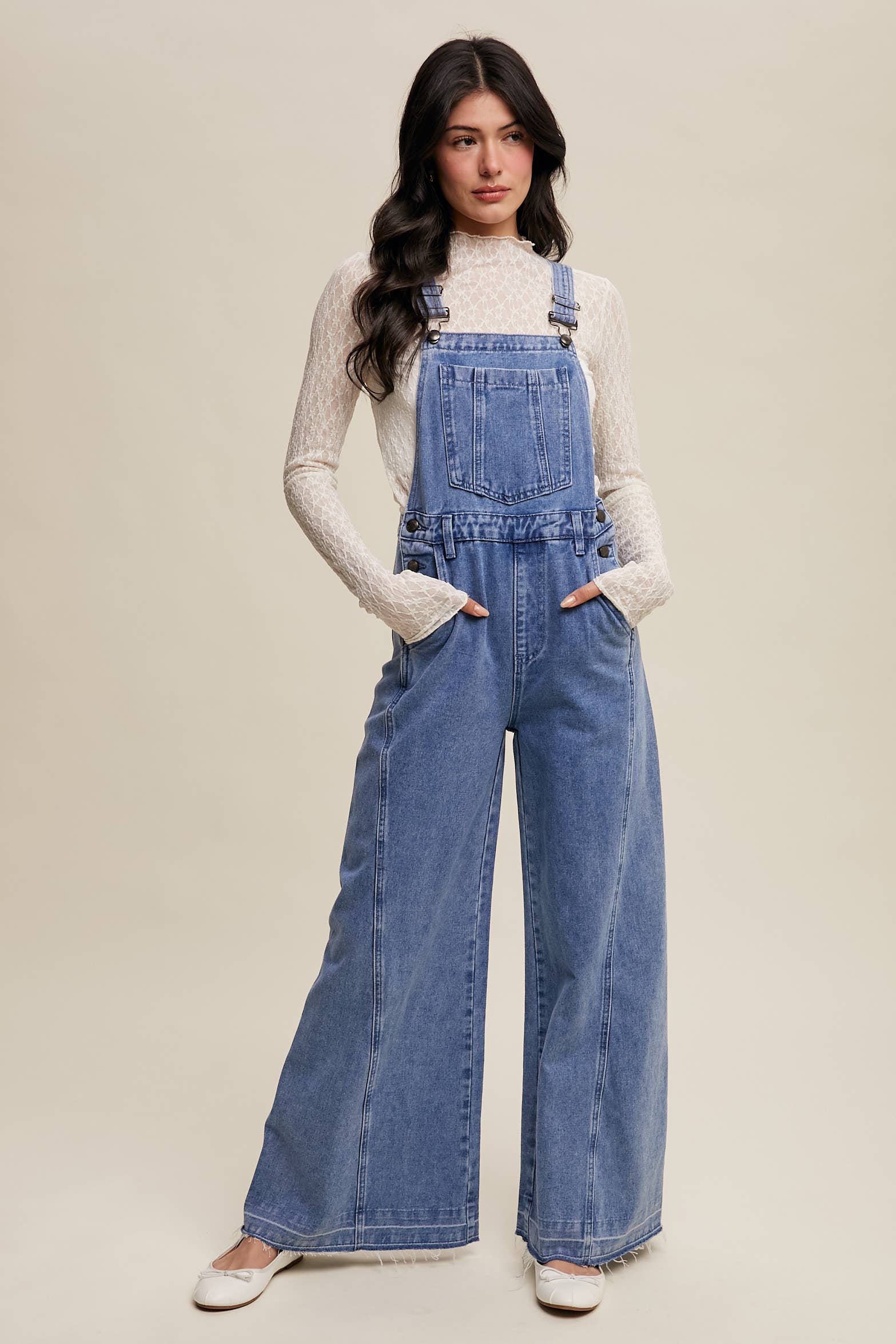 Light-wash wide-leg denim overalls with adjustable straps, front bib pocket, side buttons, and slant pockets, finished with a raw hem and layered over a cream long-sleeve top.