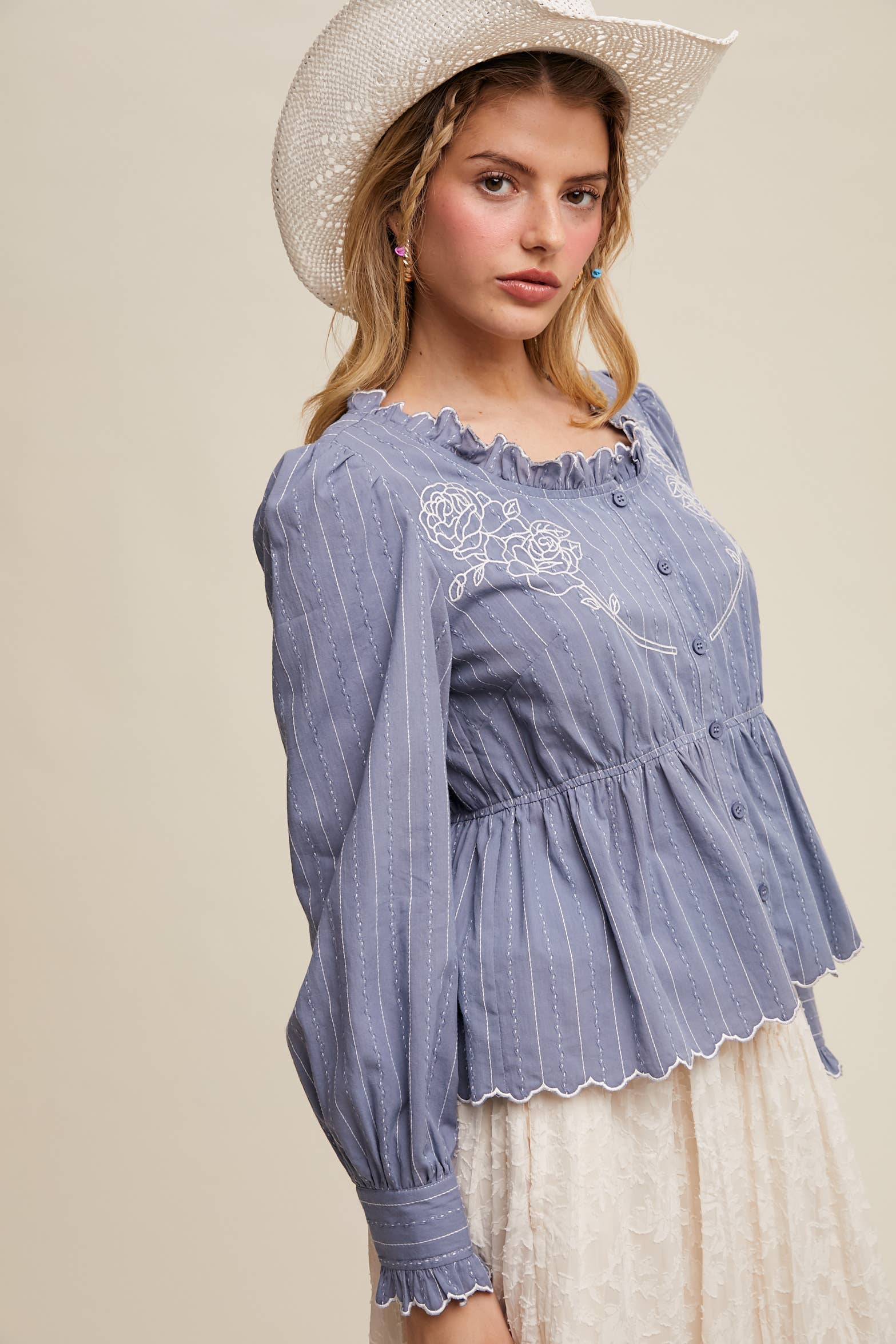 Side view- Dusty blue pinstripe blouse with ruffled neckline, white rose embroidery, puff long sleeves, button front, and scalloped peplum hem—styled with a white lace skirt and cowboy hat.