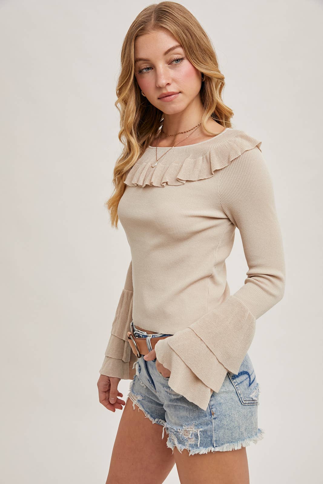 Beige ribbed sweater with ruffle neckline and tiered bell sleeves, paired with distressed denim shorts and a tan belt.