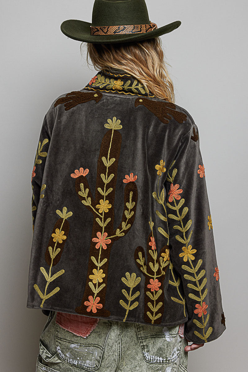 A person shown from the back wearing a dark brown velvet jacket with embroidered cactus and floral patterns in yellow, orange, and green, paired with a dark green cowboy hat and distressed denim jeans.